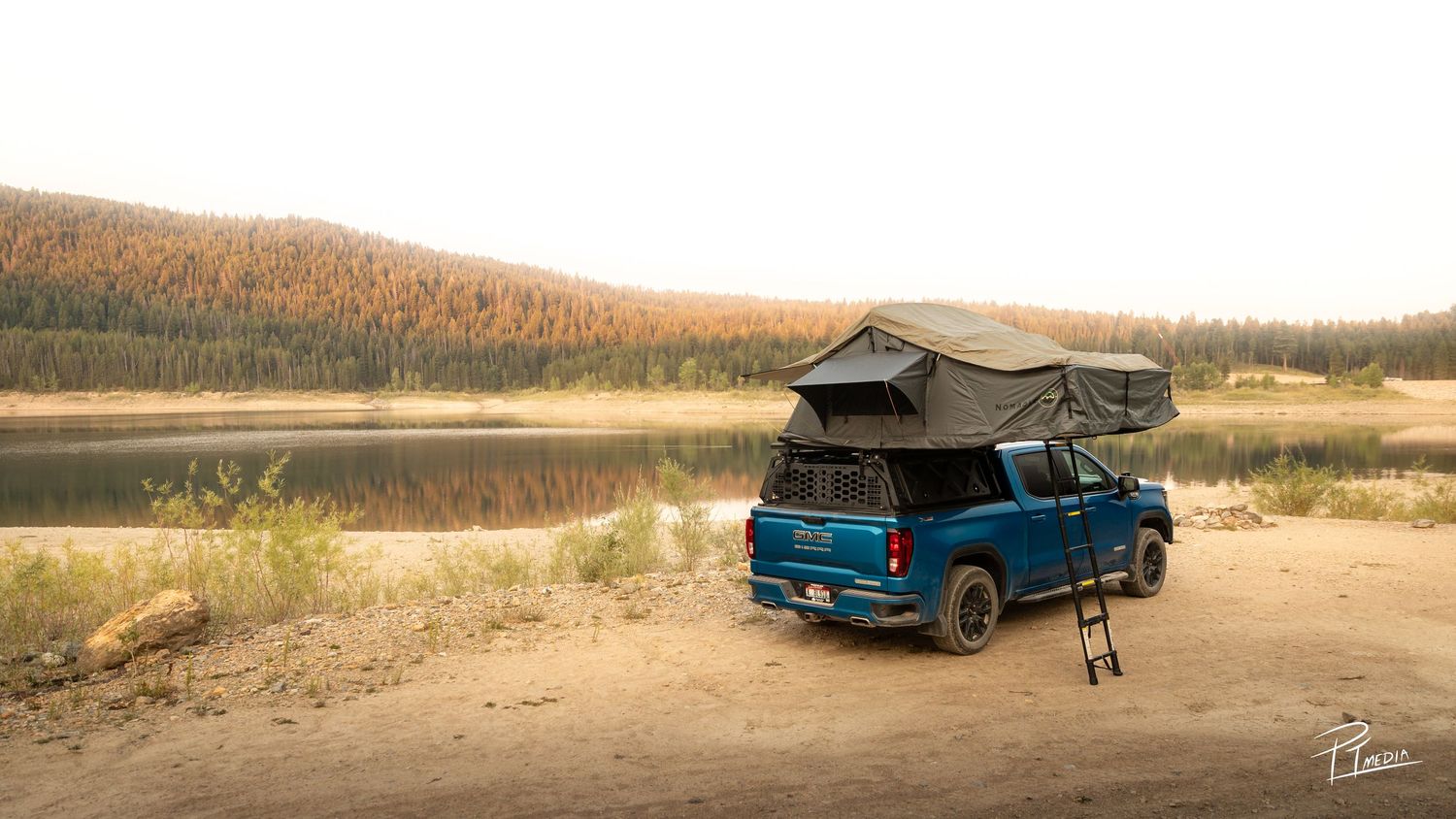 A panoramic view of a blue pickup truck with a rooftop tent camping setup parked near a scenic lake at sunset.