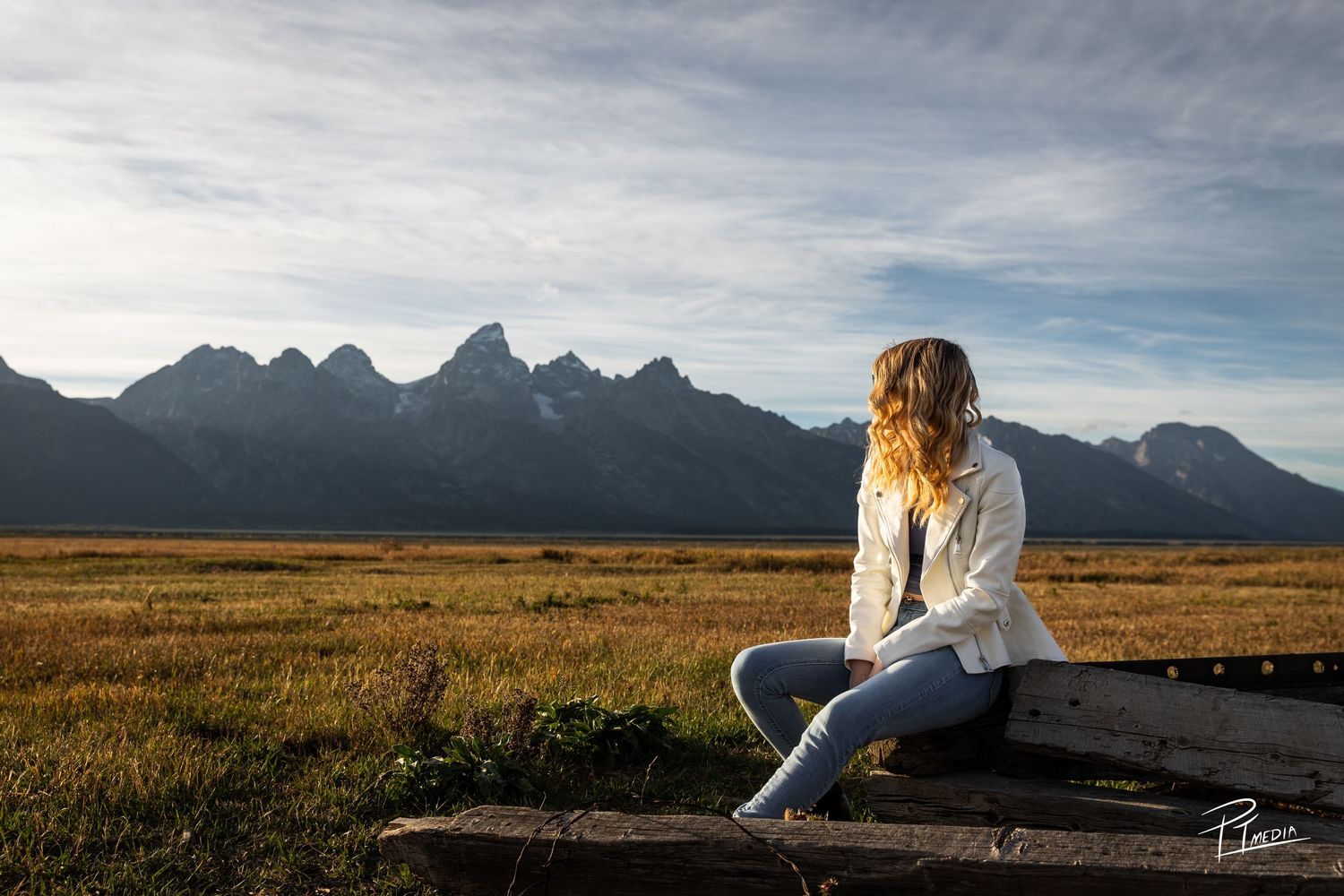 A person sits on a wooden bench overlooking a vast prairie landscape with mountains in the distance at sunset.