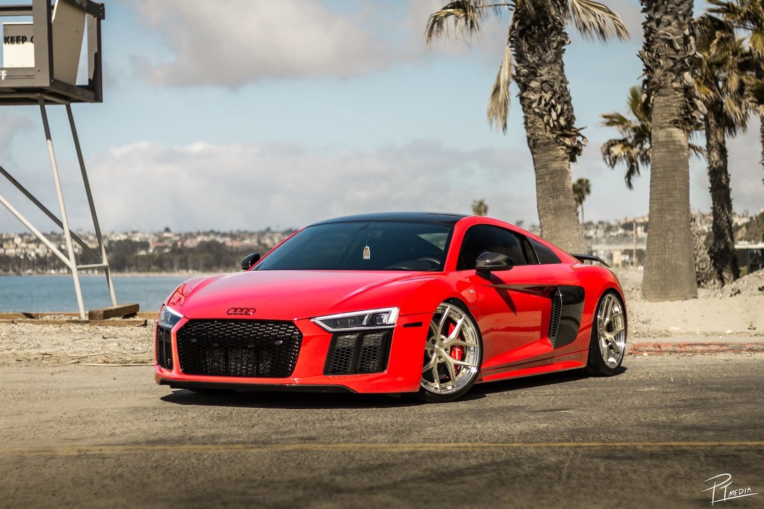 Red Audi R8 sports cars parked in sequence along a beachfront with palm trees and ocean views in the background.