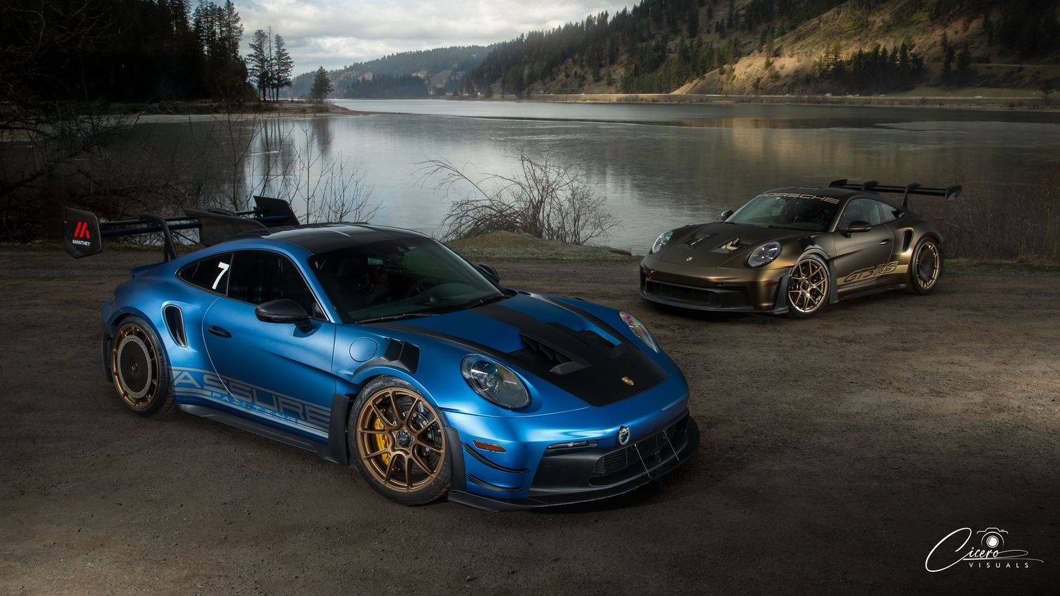 Two sports cars, a blue Porsche GT3 RS and a bronze-colored vehicle, parked by a serene lake with mountains.