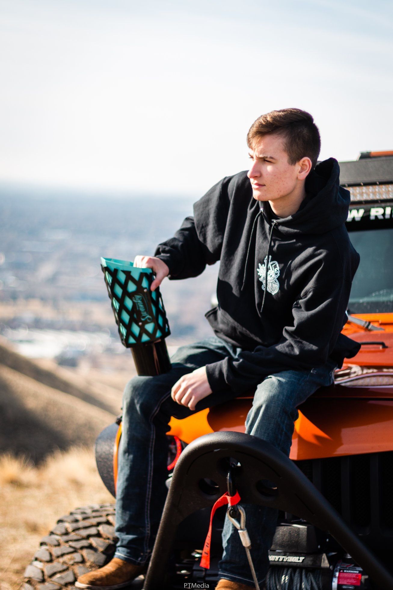 Person in black hoodie sits on orange off-road vehicle holding turquoise drink cup against scenic mountain backdrop.