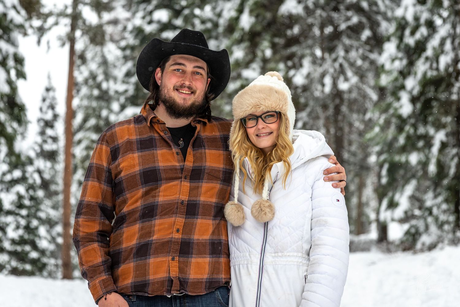 A couple poses together in a snowy winter forest setting wearing plaid flannel and white winter clothing.