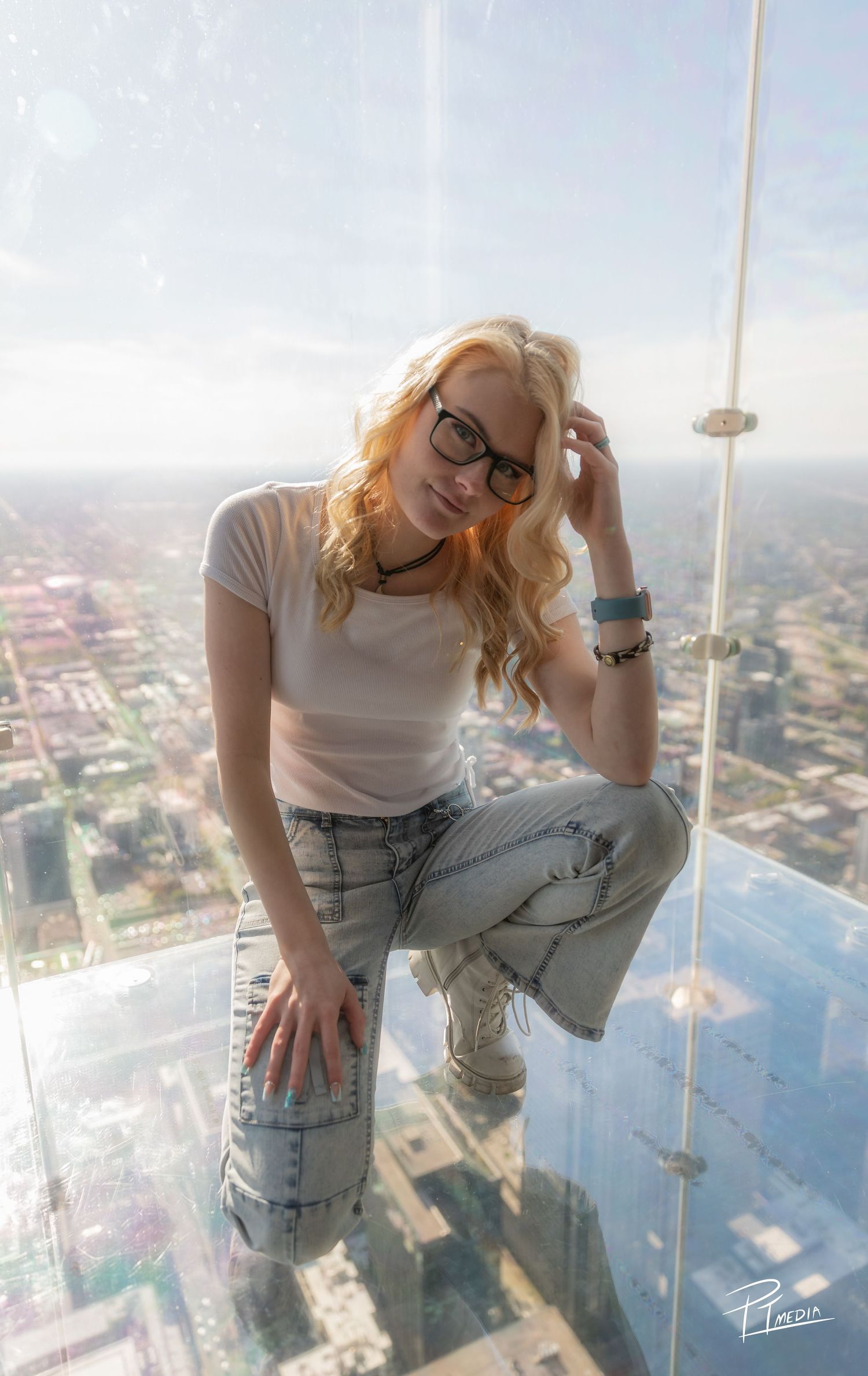 Person in ripped jeans and glasses poses playfully on glass observation deck overlooking cityscape from high above.