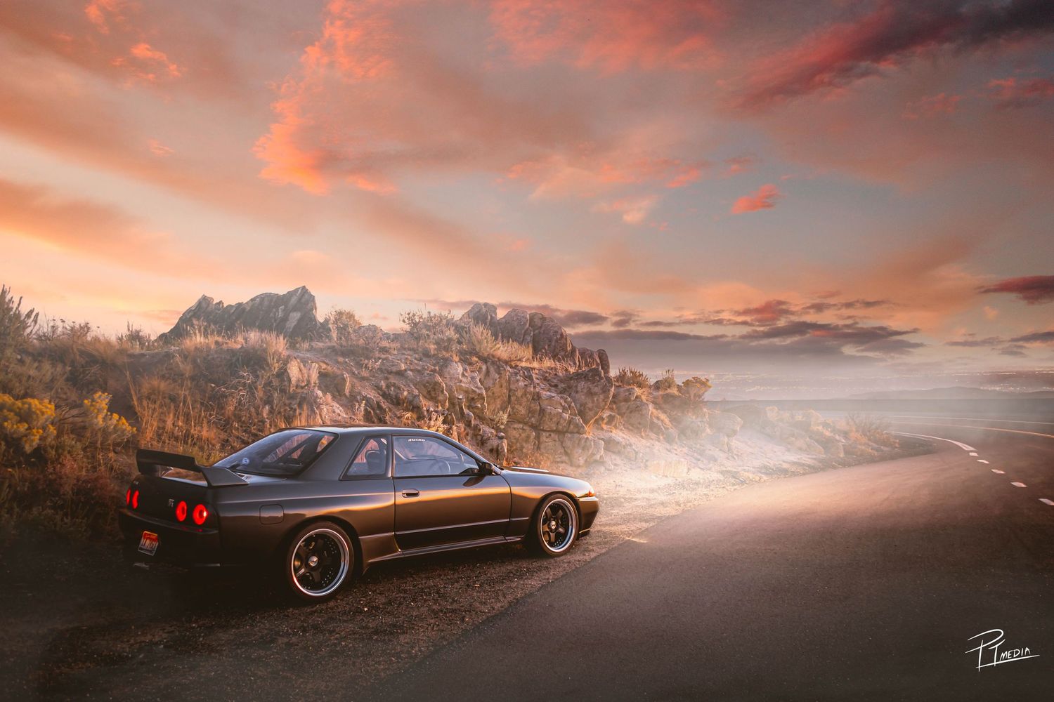 Black Nissan Skyline GTR parked on mountain road during dramatic sunset with pink and orange clouds in the sky.