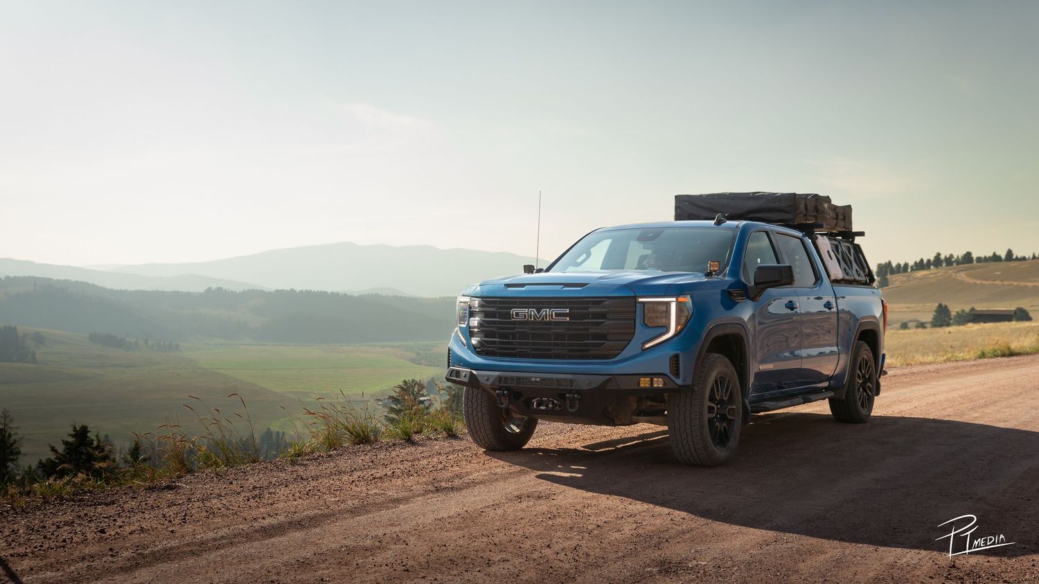 A blue GMC Sierra AT4X pickup truck driving on a rugged mountain trail during sunset with panoramic landscape views.