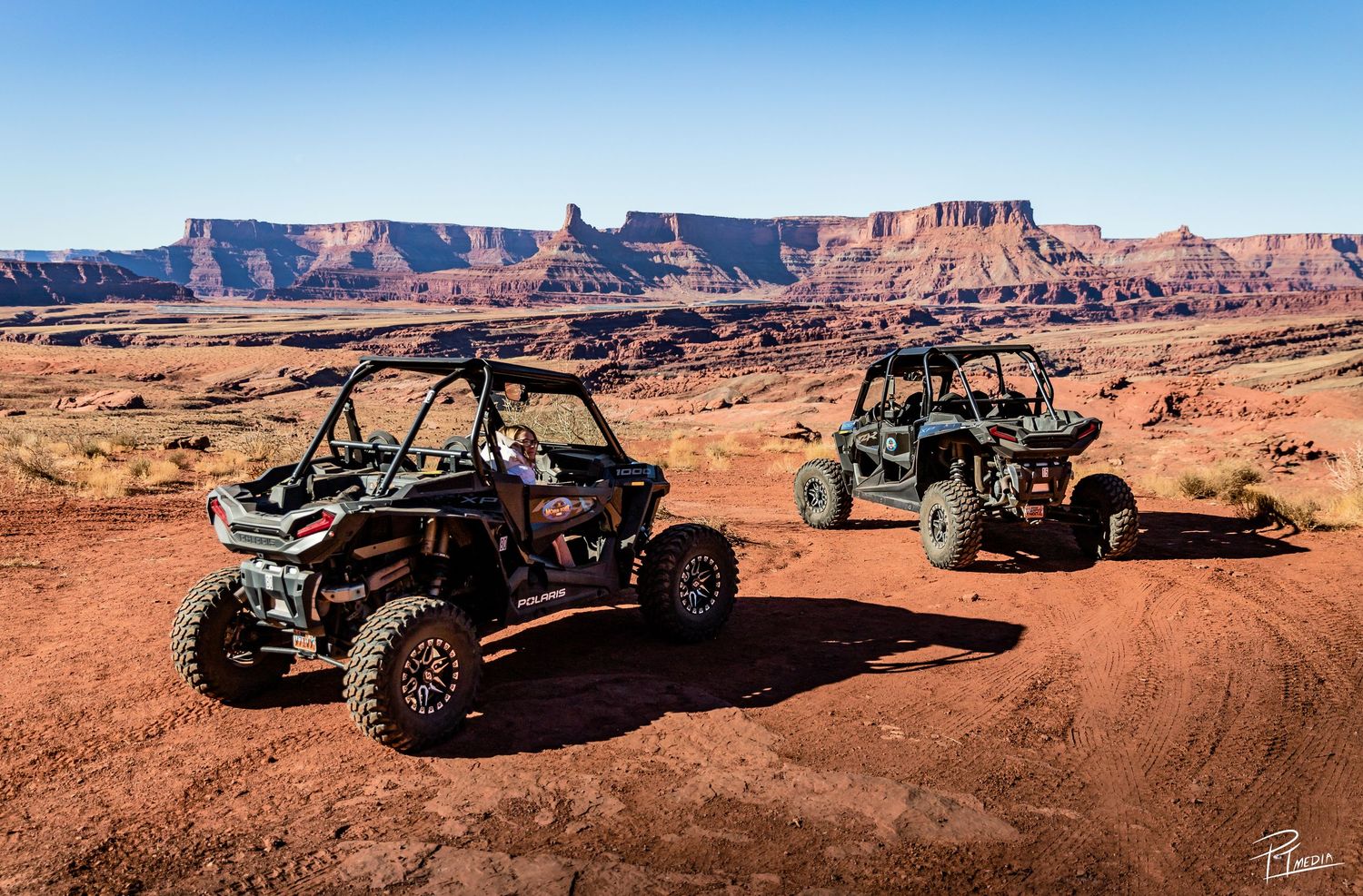 Off-road vehicles traverse the rugged red rock terrain of Moab with dramatic desert mesas in the background.