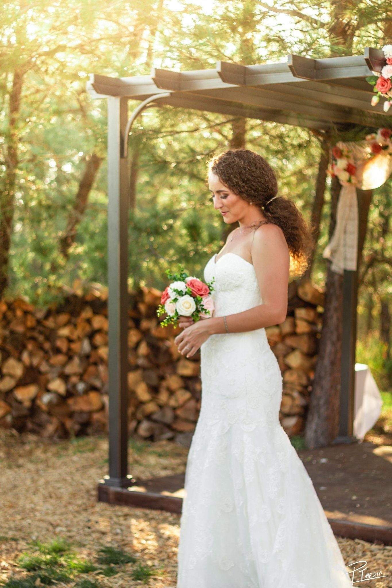 A white mermaid-style wedding dress glows in golden sunlight beneath a wooden pergola surrounded by trees.