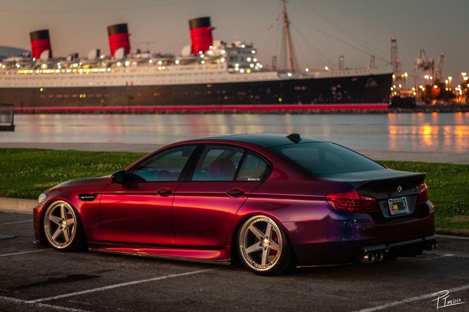 A lowered red BMW M5 parked at night by the water with the Queen Mary ship illuminated in the background.