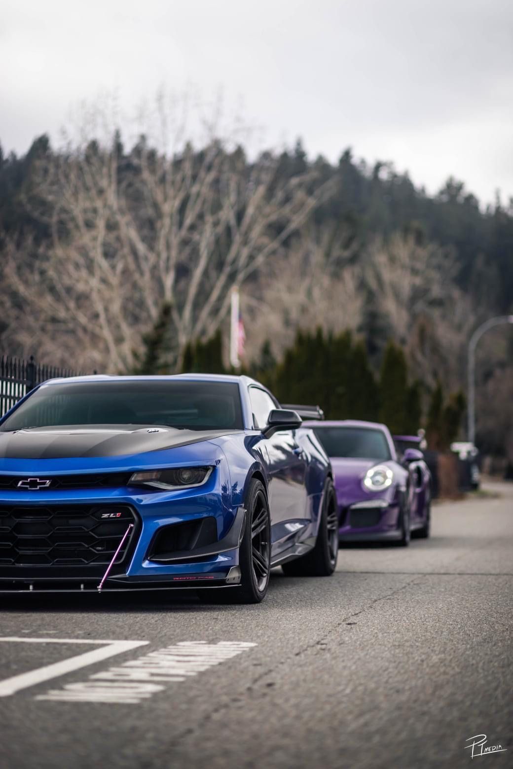 Blue Chevrolet Camaro ZL1 leads a line of performance cars parked on a street with mountains and trees in background.