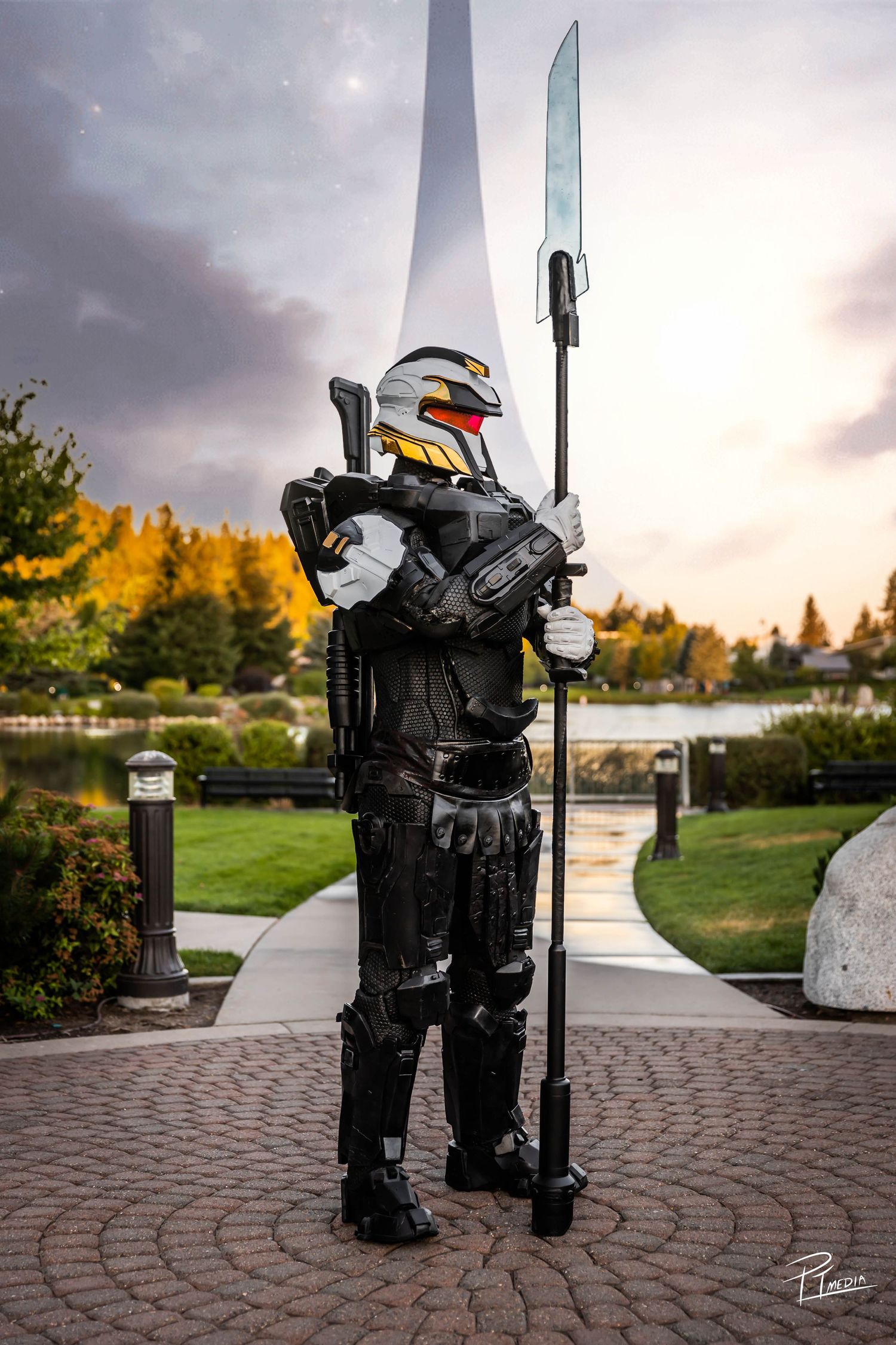 Cosplayer in futuristic black armor and colorful helmet poses with spear on brick pathway at sunset.