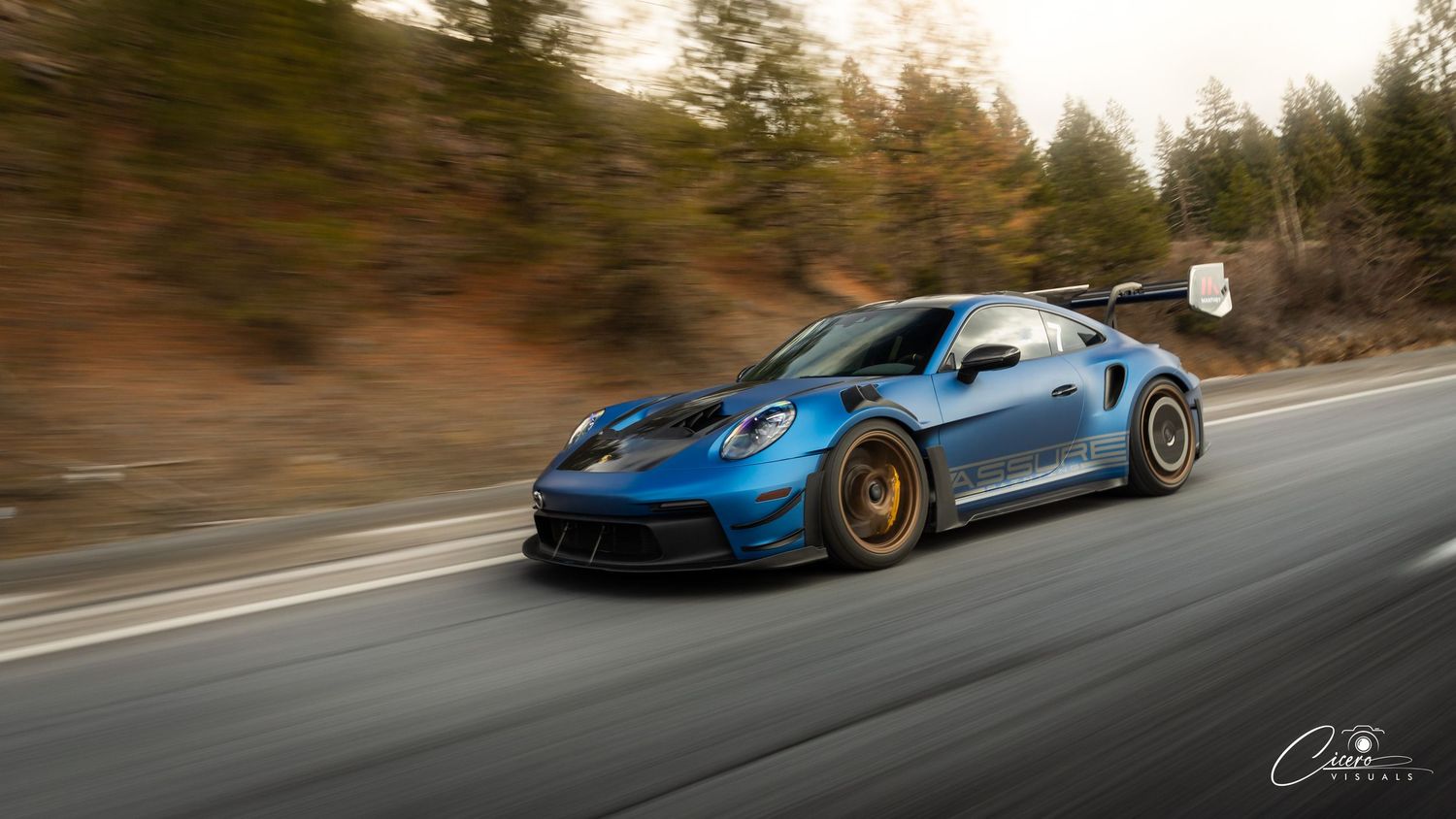 A blue Porsche 911 GT3 RS sports car speeds along a mountain road with motion blur effect.