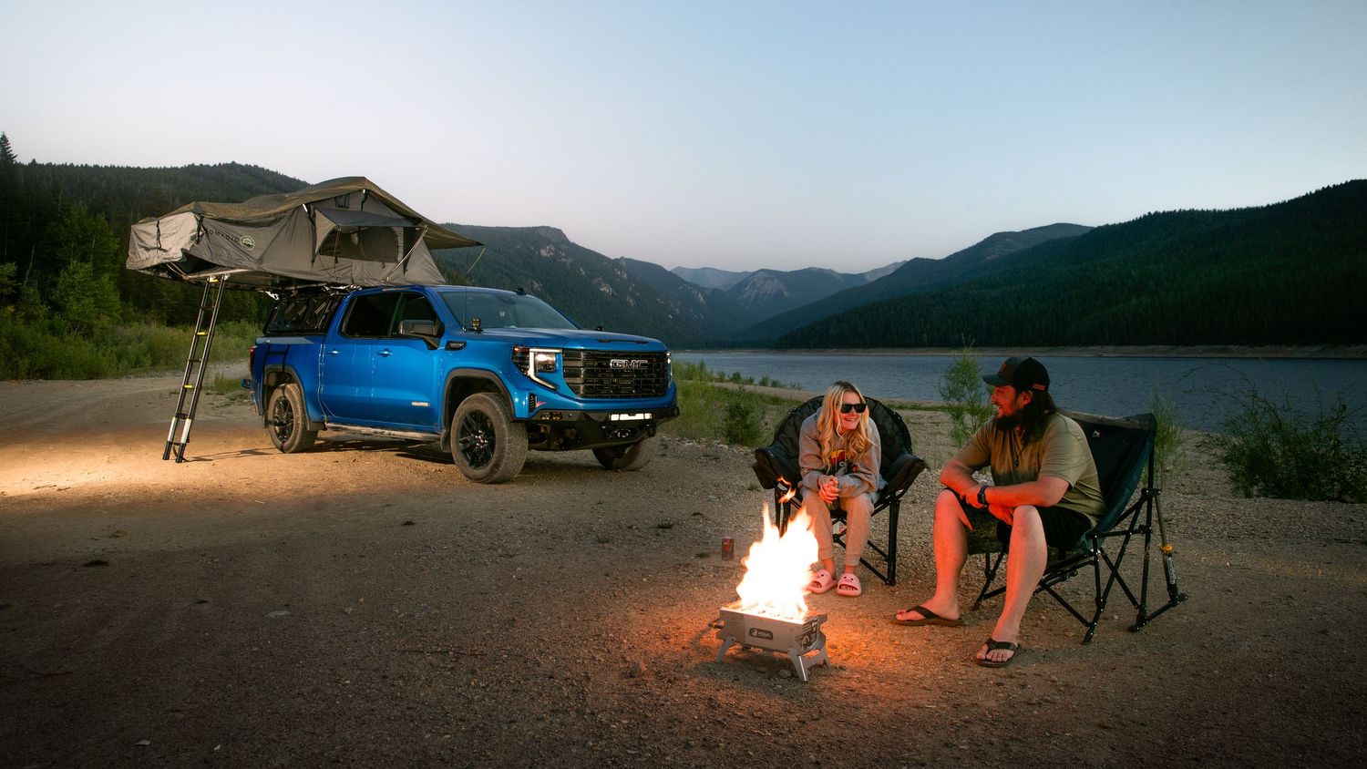 A panoramic mountain landscape at sunset with campfire glowing against rugged terrain and twilight sky.