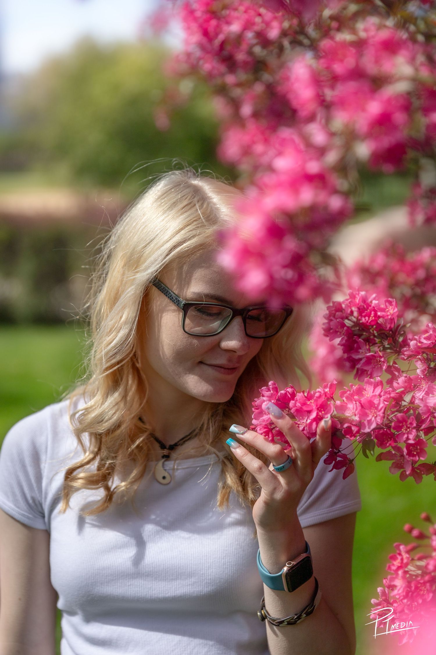 A person in a white t-shirt smells vibrant pink cherry blossoms on a spring day in a garden setting.