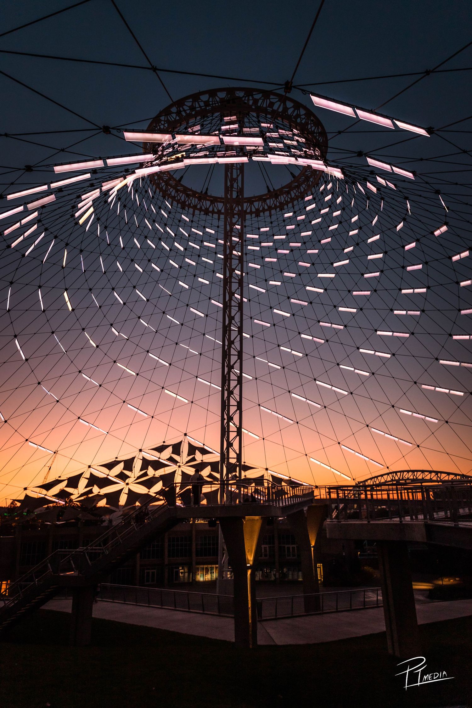 Geometric light installation with pink-tinted panels creates striking patterns against sunset sky at outdoor venue.
