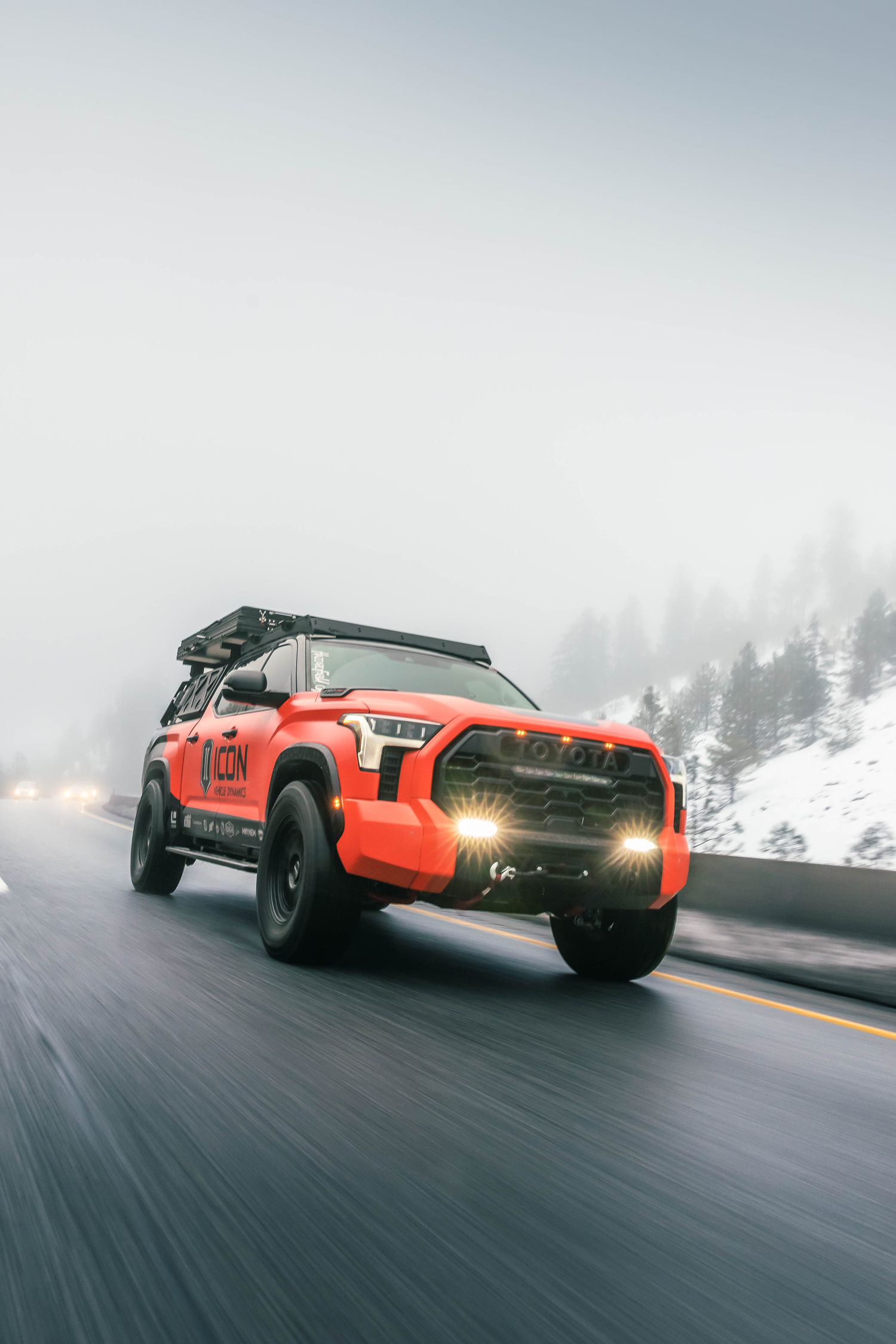 Orange off-road vehicle speeding through snowy mountain roads with fog in the background.