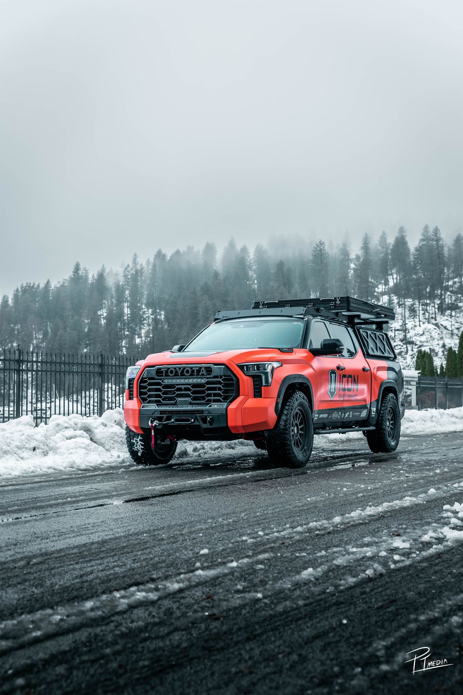 Orange off-road vehicle driving through snowy mountain terrain with evergreen trees in misty background.