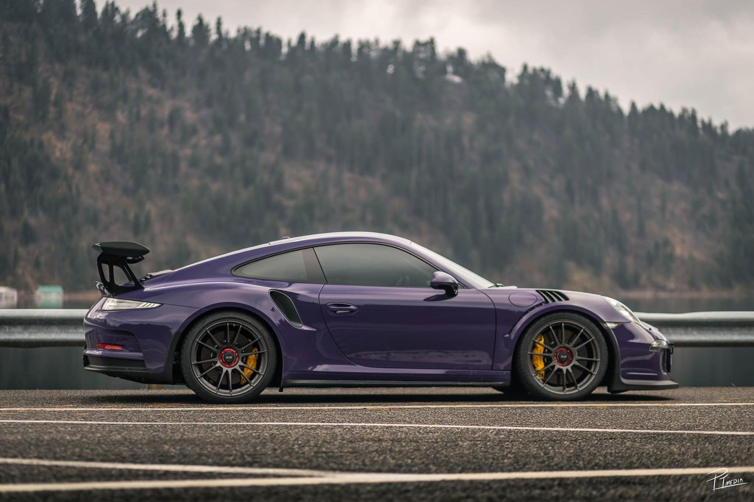 Purple McLaren supercar photographed in motion on a racetrack with forested mountains in the background.