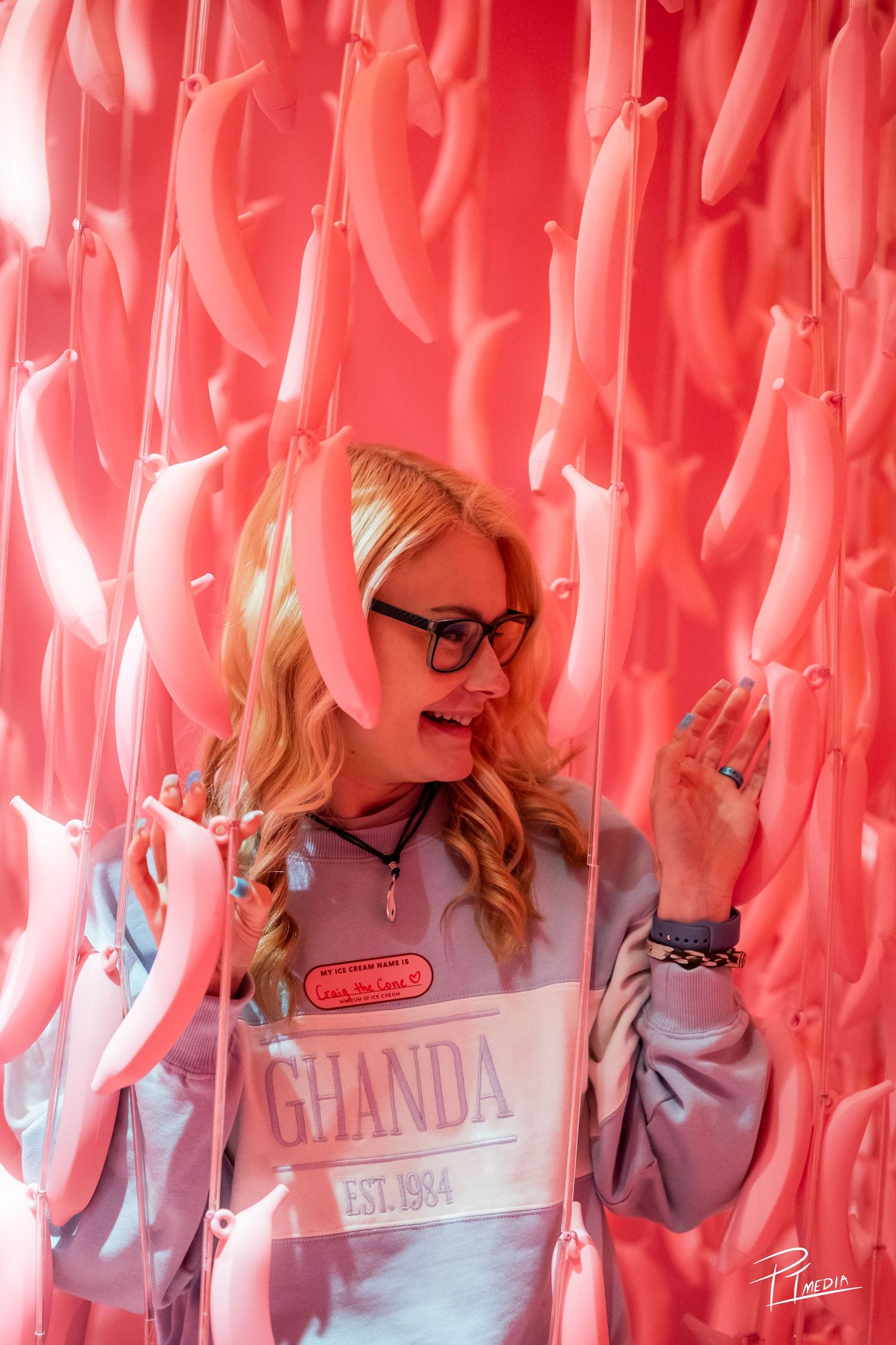 A person in a gray sweater stands against a vibrant pink backdrop with hanging pink ribbons or streamers.