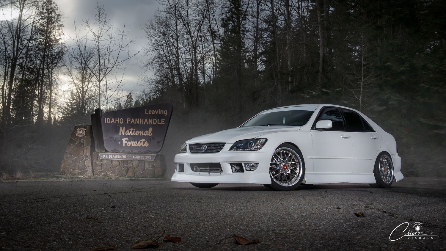 A white modified Lexus IS300 with custom wheels parked in front of a forest entrance sign at dusk.