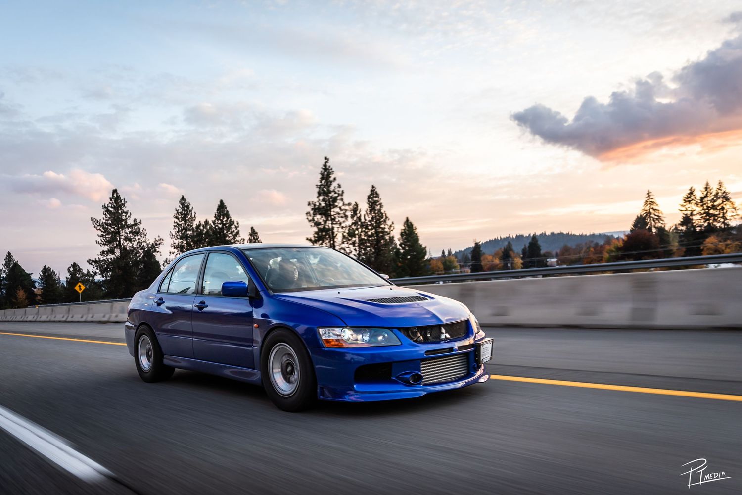A blue Nissan Skyline GTR R34 photographed at sunset against scenic mountain and coastal backgrounds.
