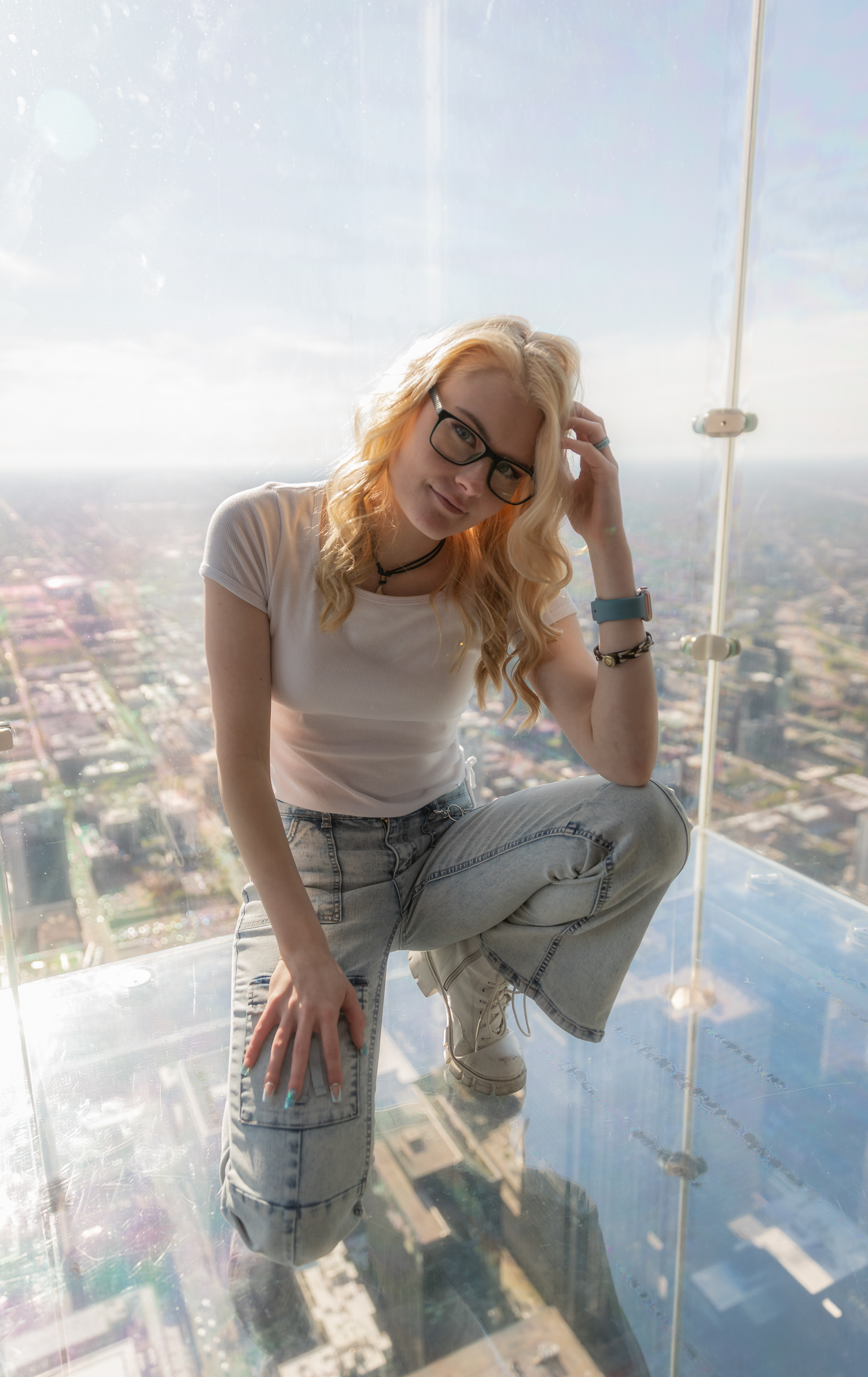 A visitor crouches on a glass skyscraper viewing platform overlooking a city below.