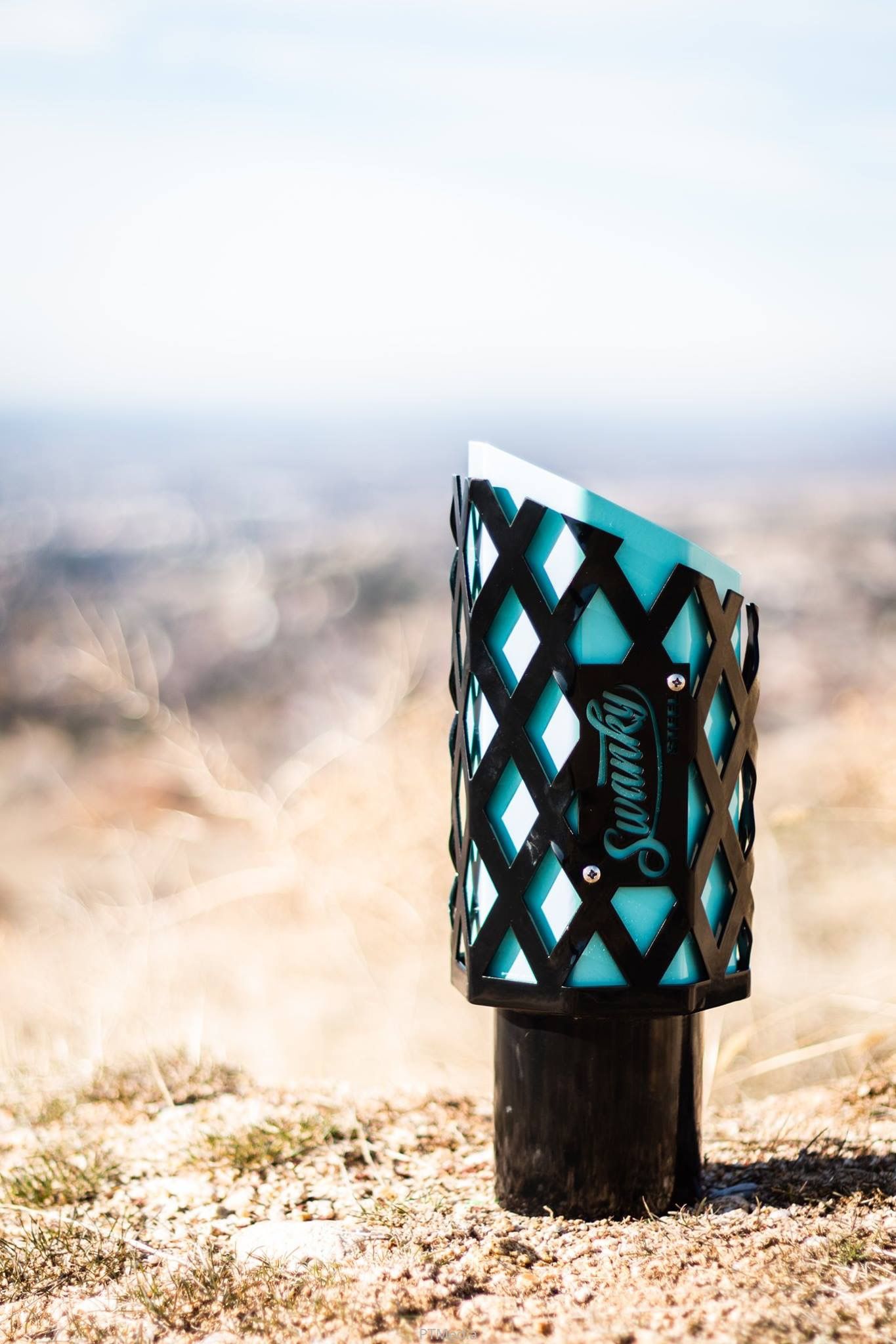 A black and turquoise decorative vape drip tip with diamond pattern design sits on sandy ground.