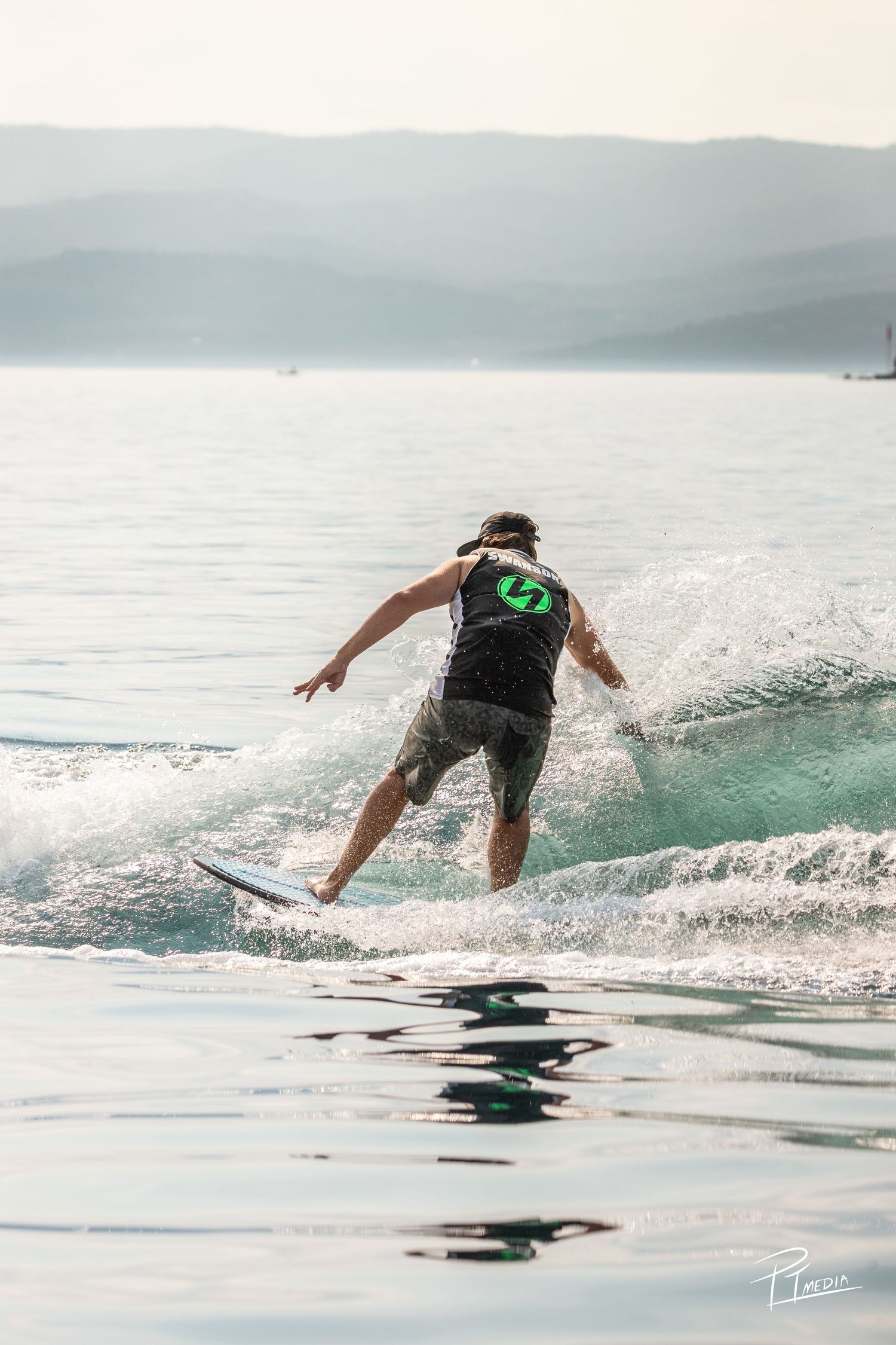 A surfer rides a wave on a surfboard in tranquil blue-green waters with misty mountains in the background.