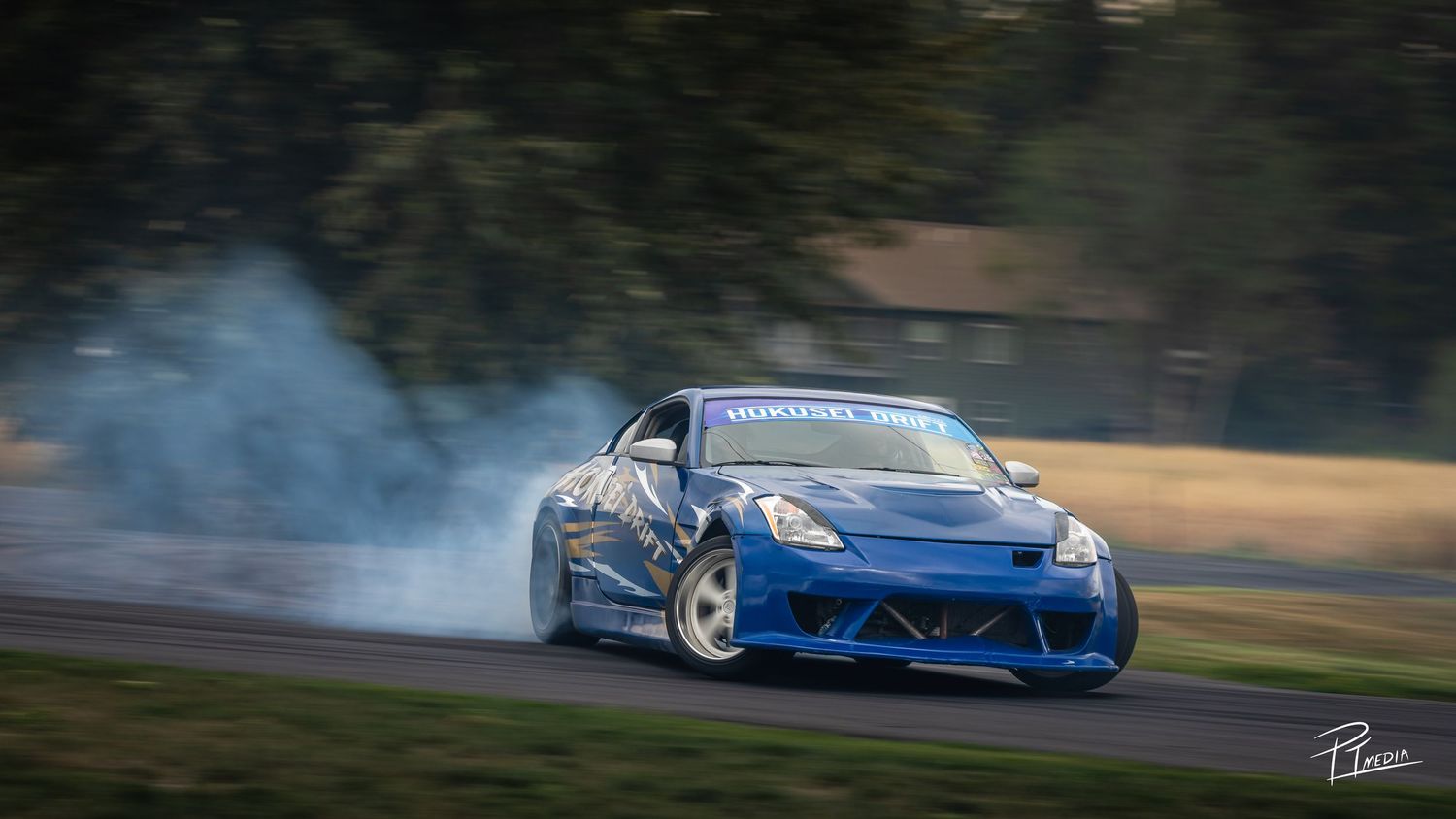 A blue sports car drifts around a track with smoke billowing from its tires during a drift competition event.