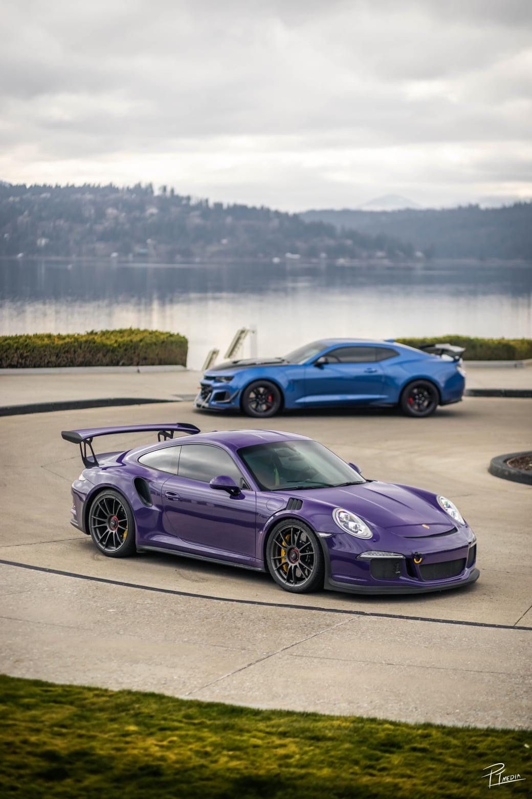 A purple Porsche 911 GT3 RS sports car parked on a waterfront with mountains and water in the background.