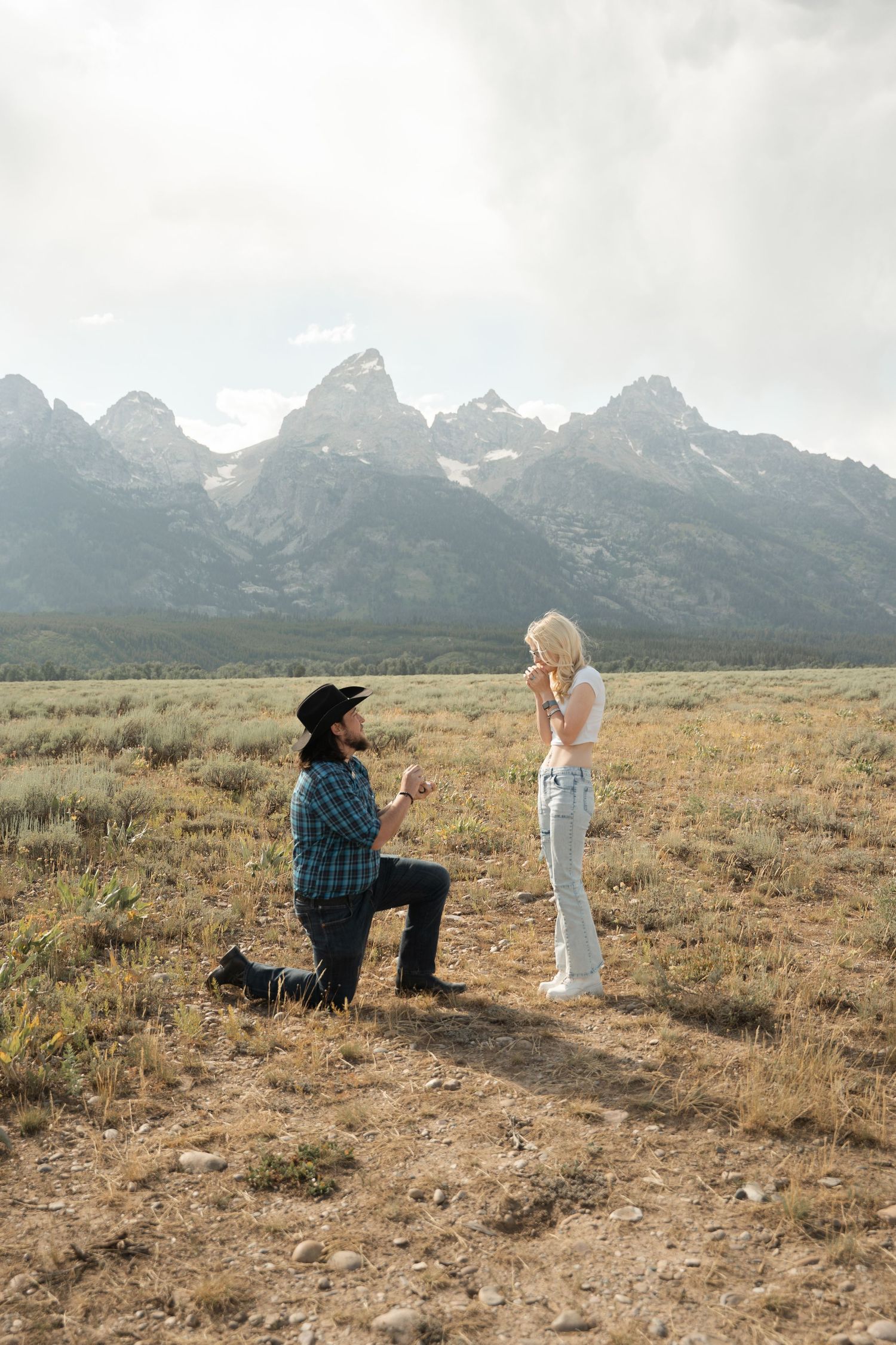 A romantic proposal moment unfolds in a vast field with dramatic mountain peaks rising in the background.