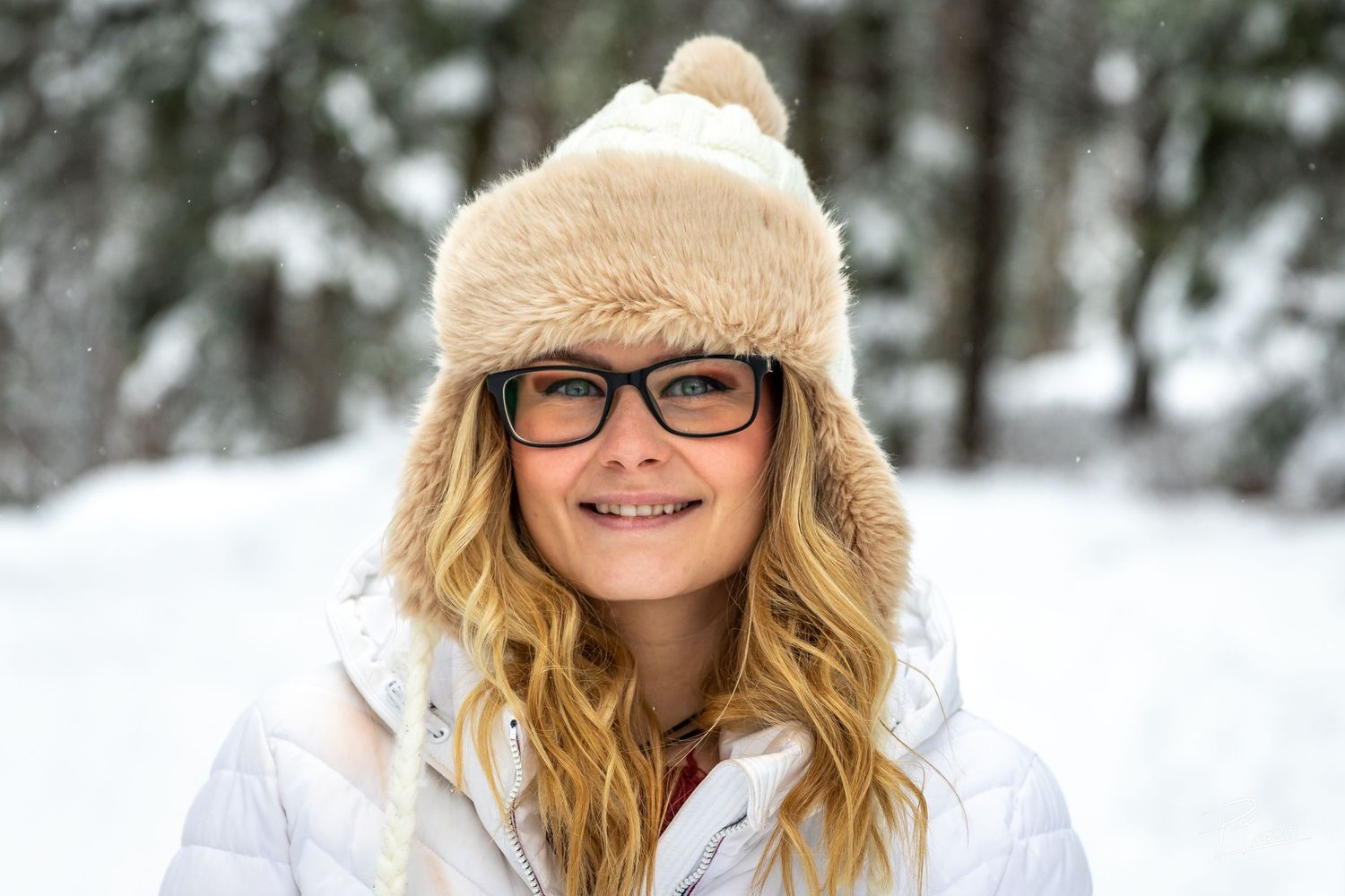 A person wearing a beige winter hat and glasses smiles while standing in a snowy winter landscape.