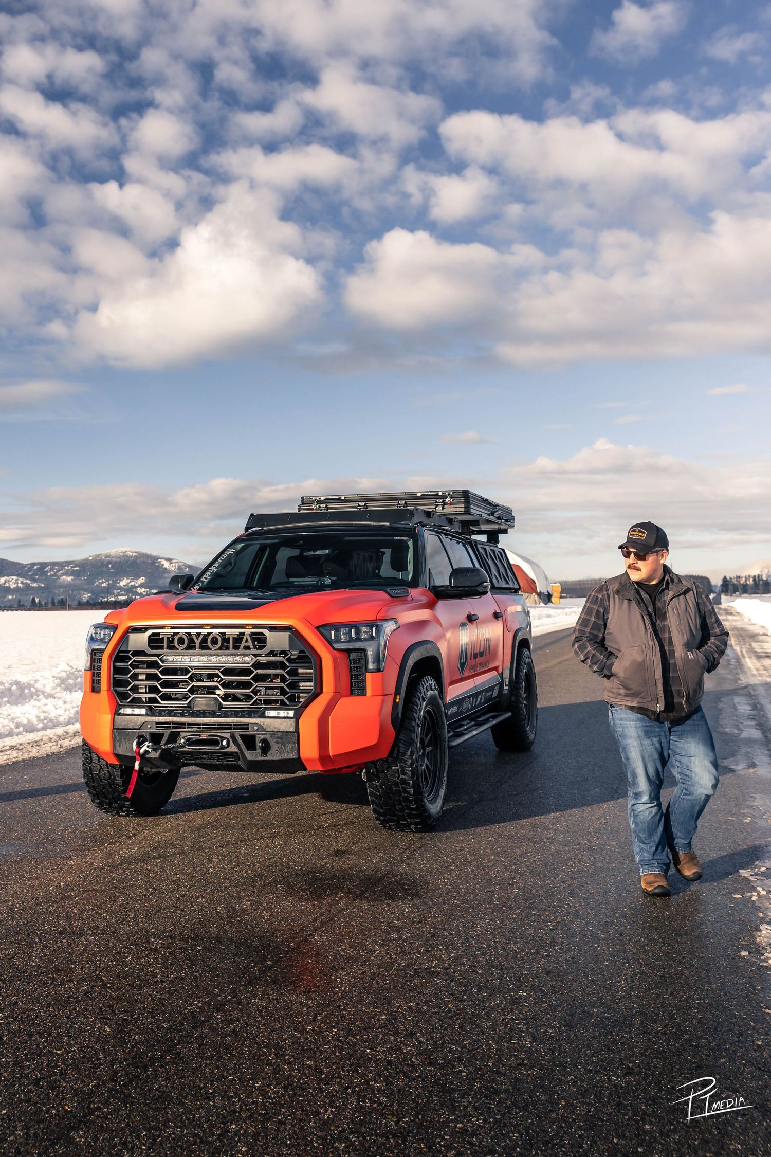 Custom orange Toyota Tundra pickup truck with off-road modifications parked on black volcanic terrain under cloudy skies.
