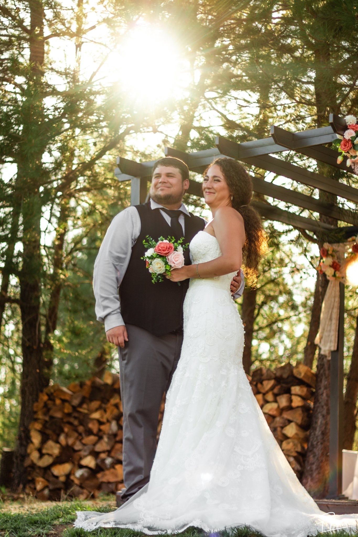 Newlywed couple shares romantic moments at sunset with stacked firewood and forest backdrop in outdoor wedding venue.