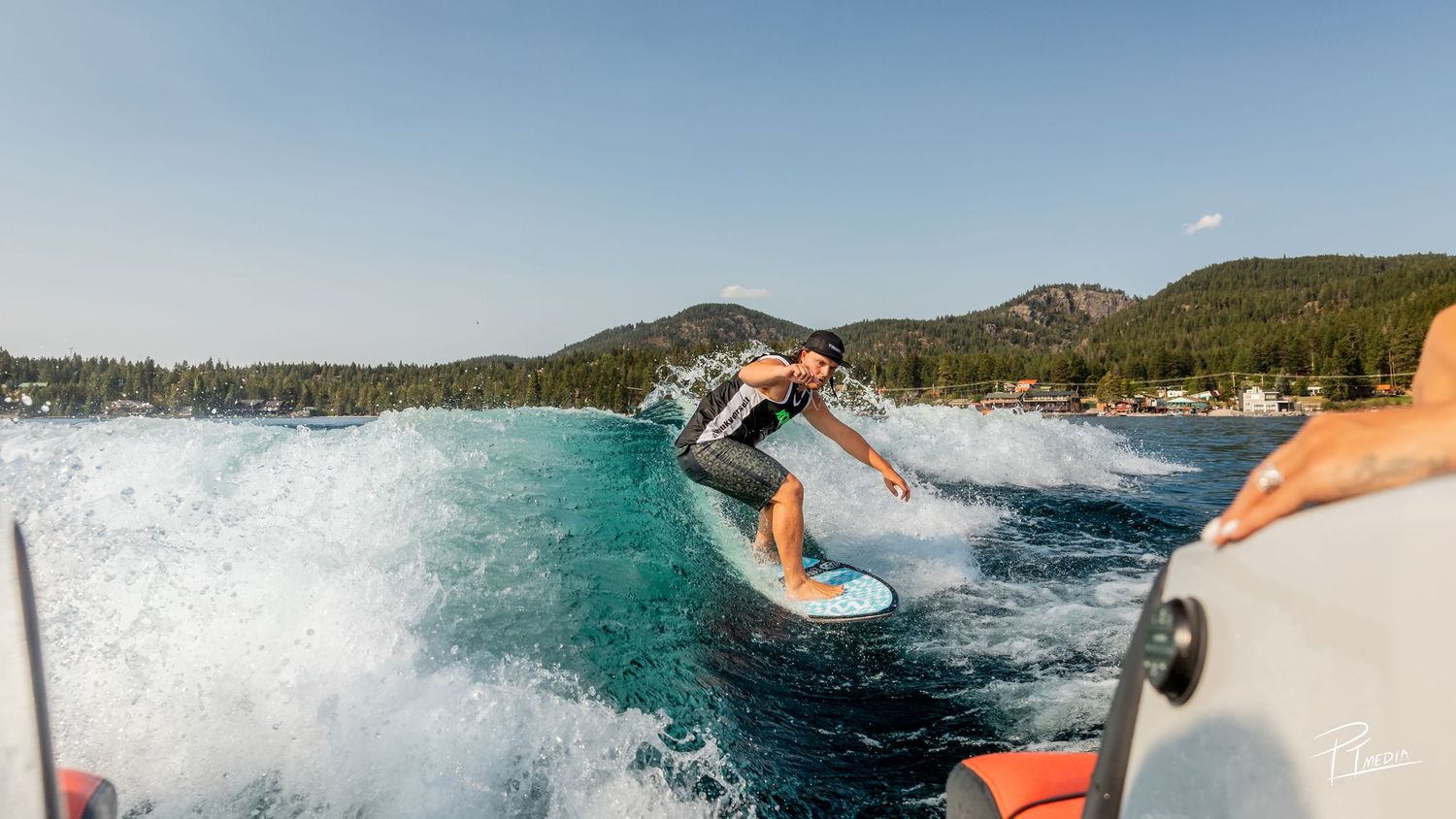 A panoramic view of surfers riding waves in a scenic bay surrounded by mountains and forest.