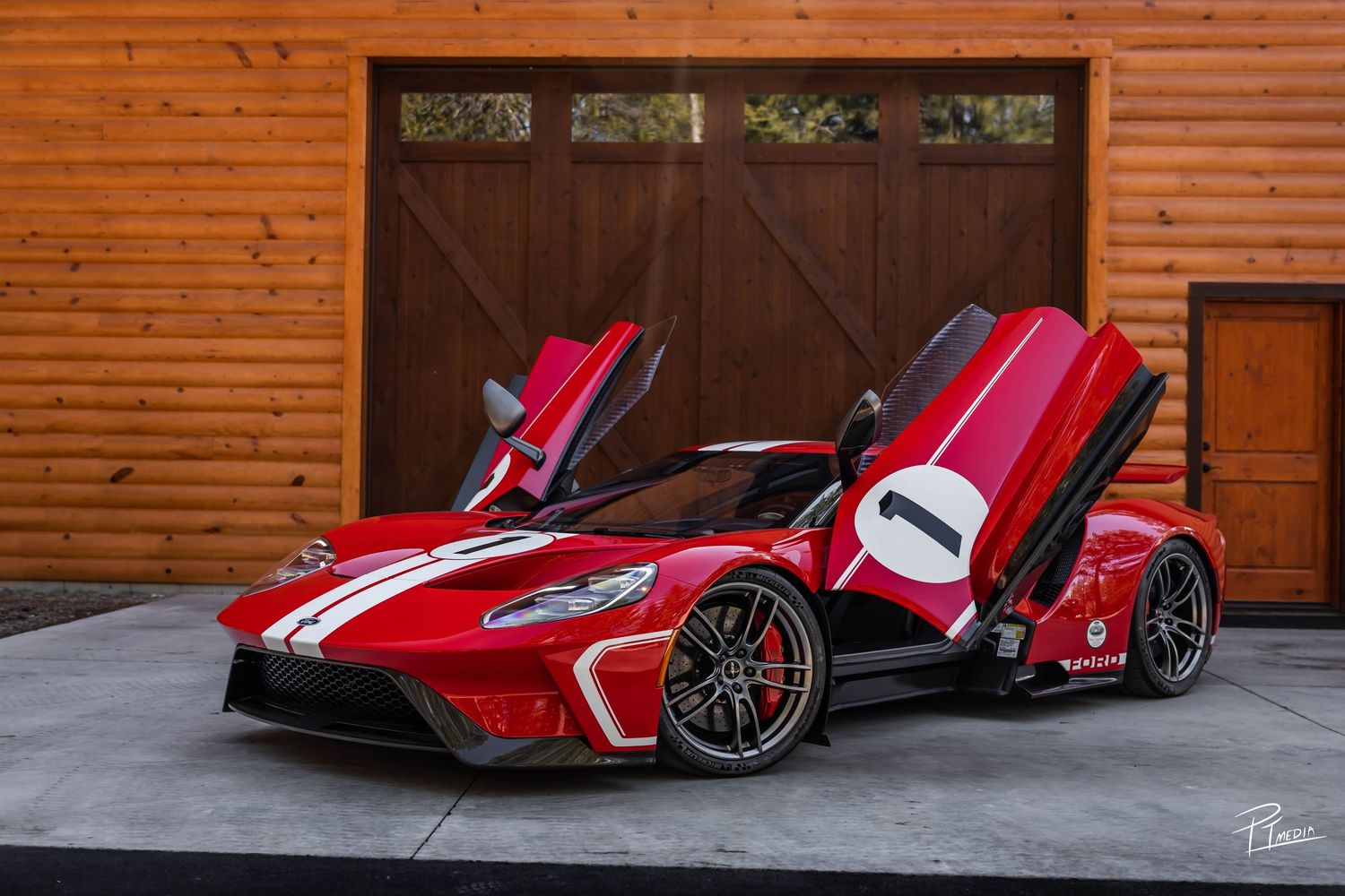A red Ford GT supercar with white racing stripes and scissor doors open, parked in front of a wooden garage door.