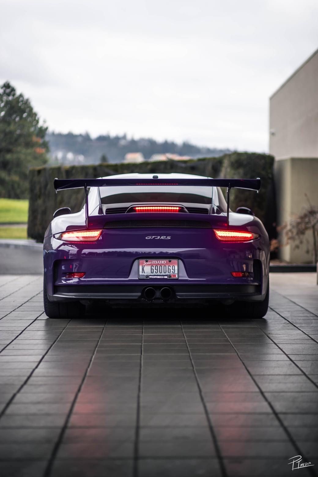 Rear view of a purple Porsche 911 GT3 RS sports car with large wing spoiler on tiled pavement at dusk.