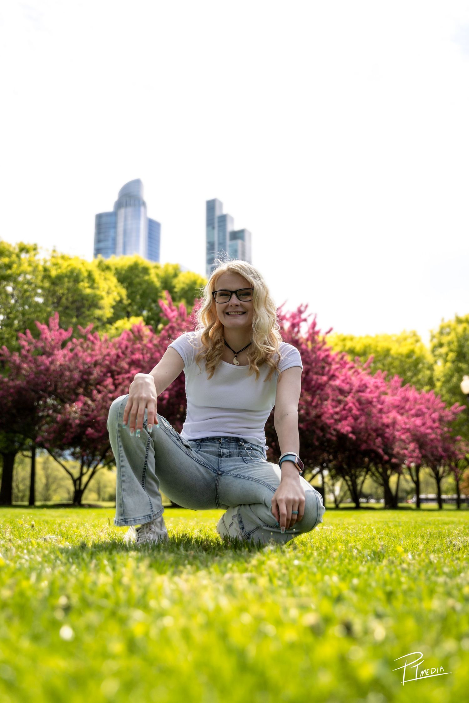 A person in jeans and sunglasses sits on lush grass with blooming cherry trees and city buildings in the background.