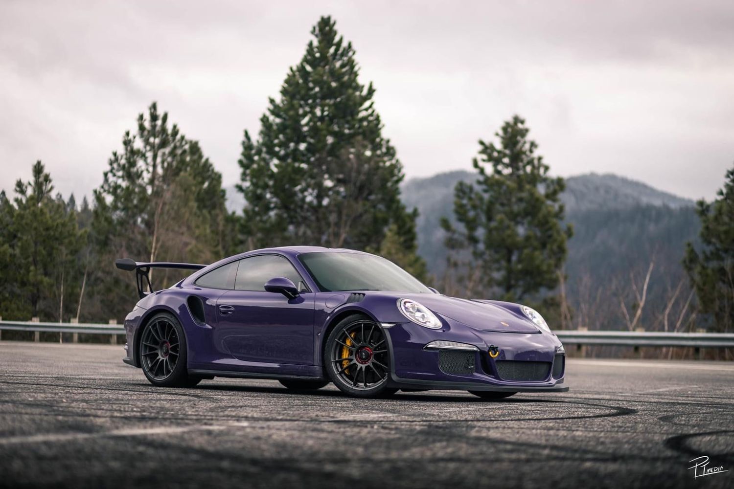 Purple Porsche 911 GT3 RS sports car parked on mountain road with evergreen trees and mountains in background.