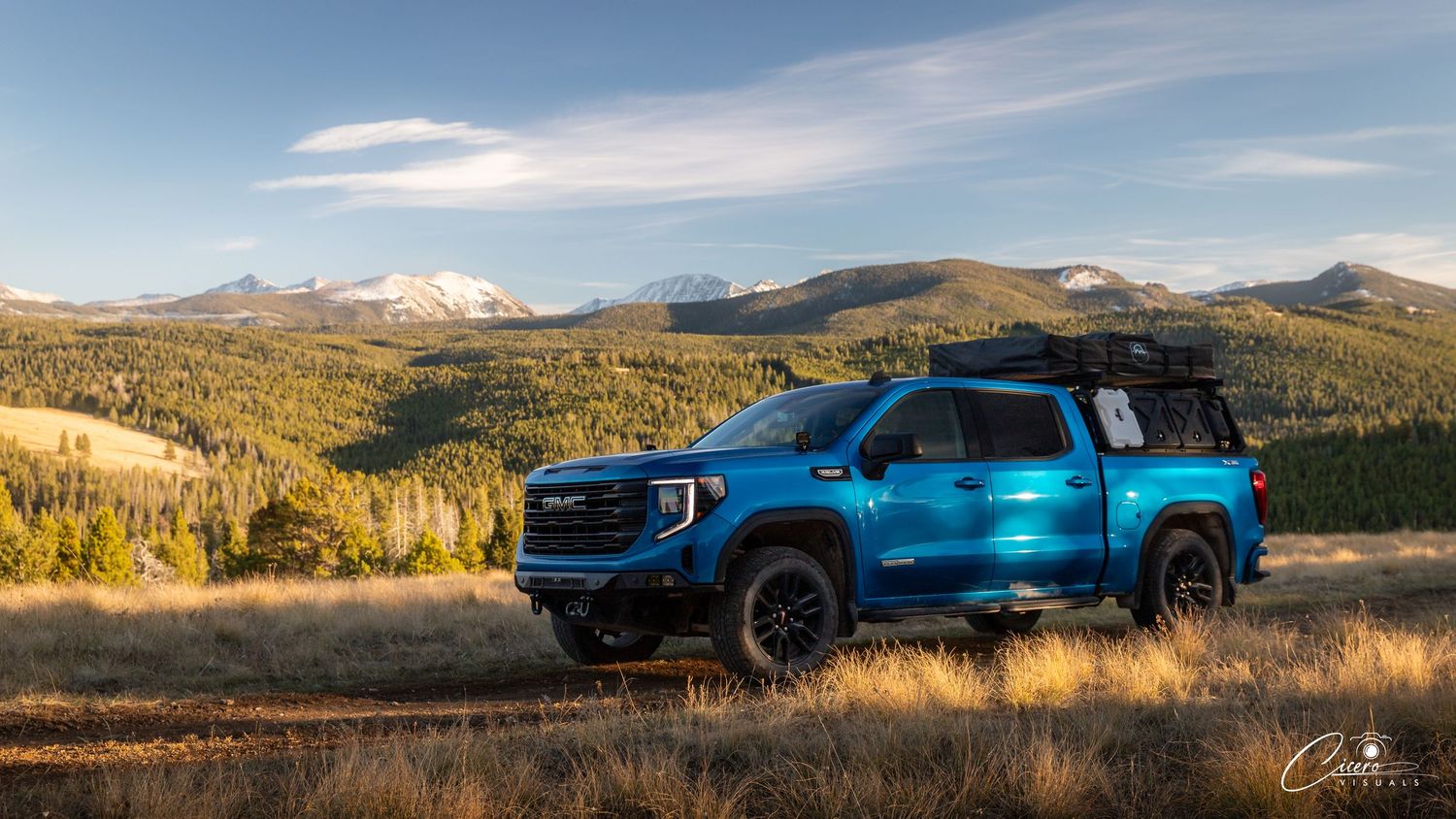 A blue GMC Sierra pickup truck parked in a scenic mountain landscape with autumn foliage and snow-capped peaks.