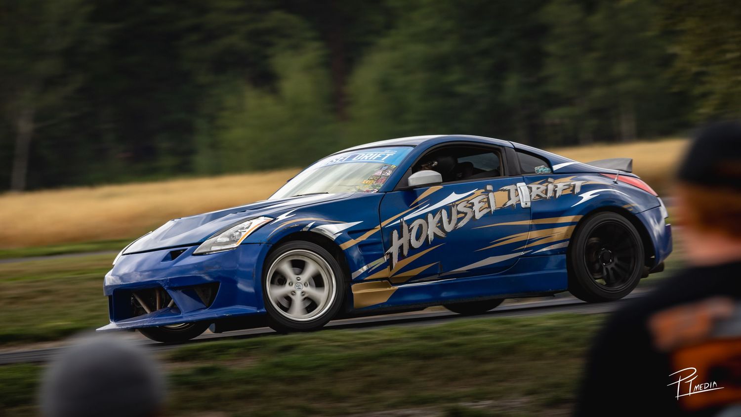 A blue sports car with racing stripes and custom modifications speeds along a racetrack during a performance event.