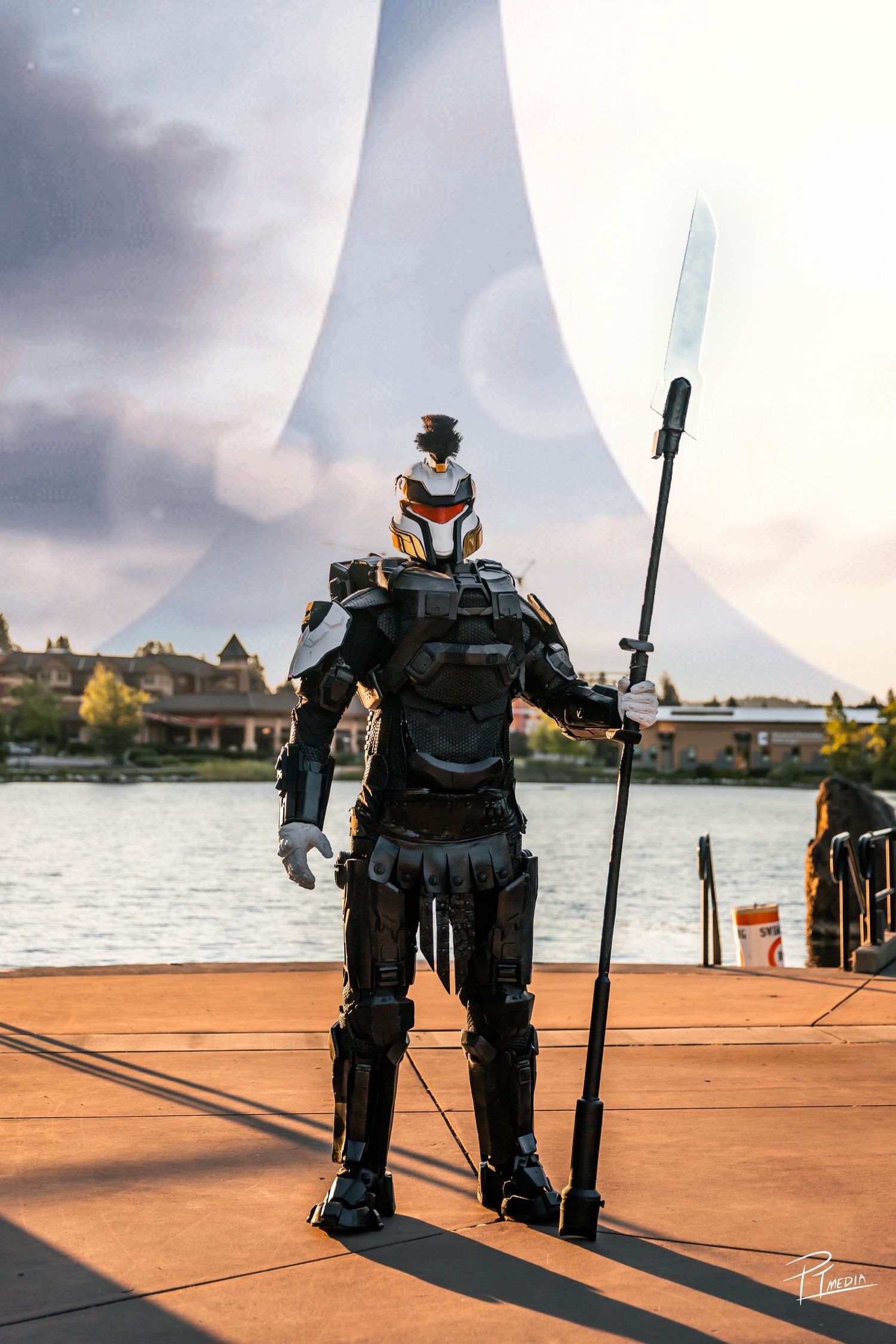 A small black robot stands on a waterfront pier against a cloudy sunset sky casting long shadows.