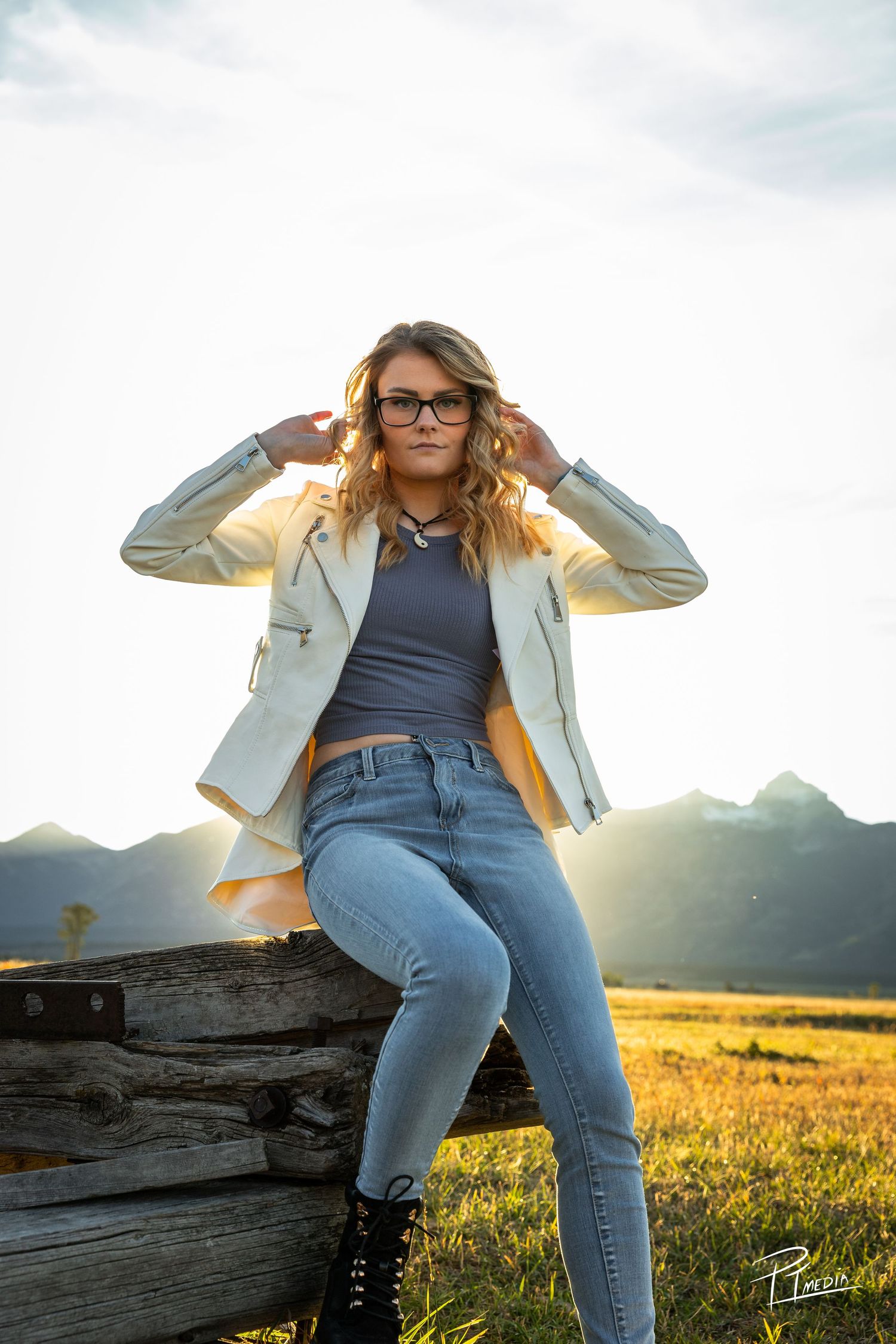 Woman in denim and beige blazer poses on wooden fence at sunset in golden field with mountains in background.