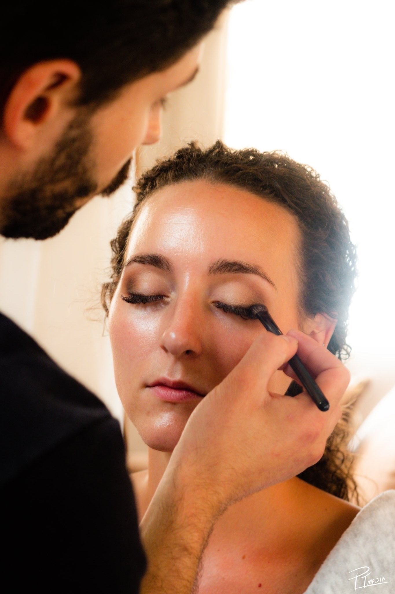 Professional makeup artist applies eye makeup with brush in warm lighting during beauty session.