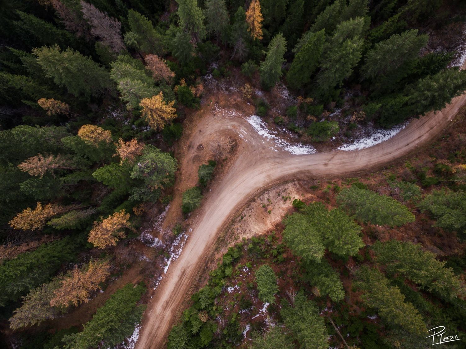 Aerial view of a winding dirt road cutting through dense forest with autumn foliage in mountain terrain.