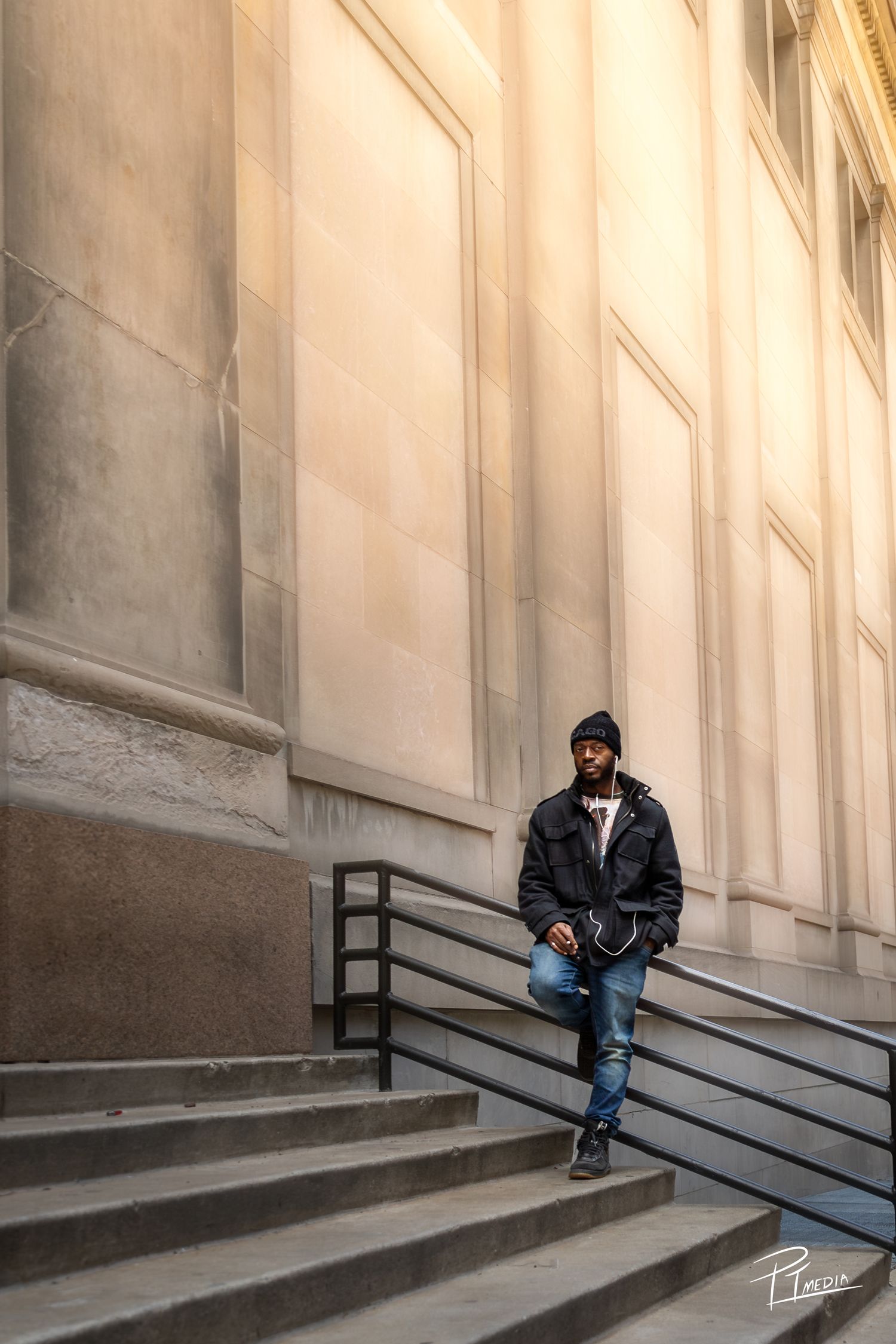 A person in dark clothing sits alone on concrete steps against a light-colored building wall in moody lighting.