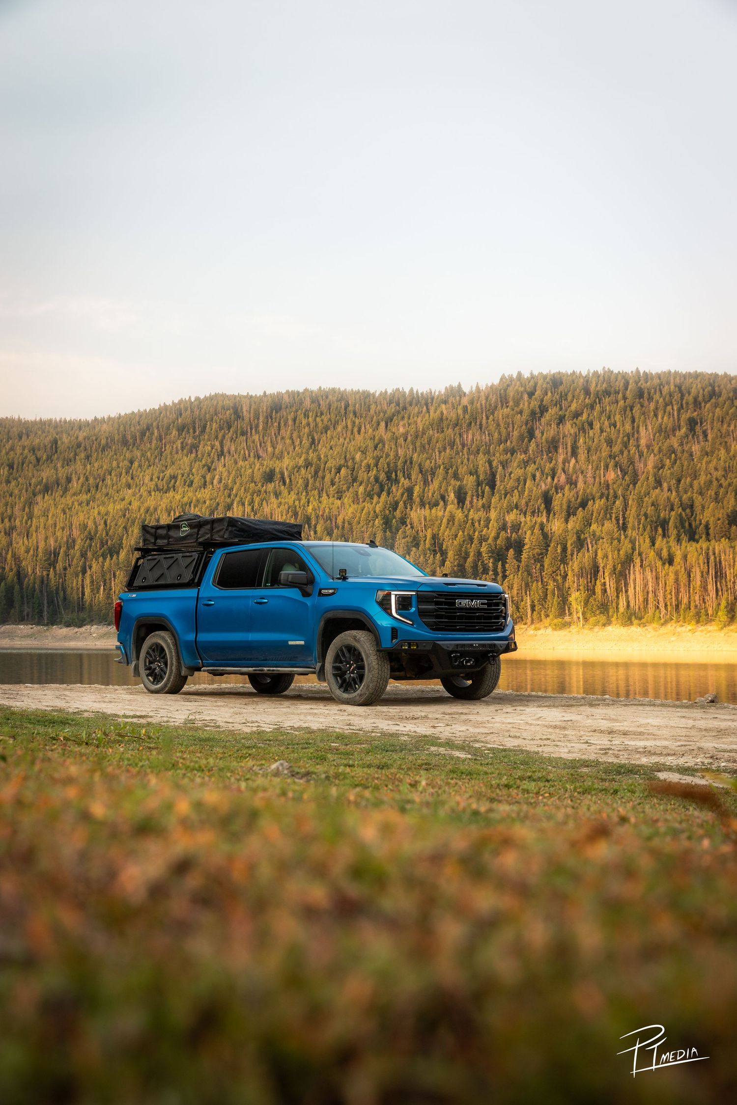 Blue Ford F-150 pickup truck parked by a lake with forested hills in the background during golden hour.