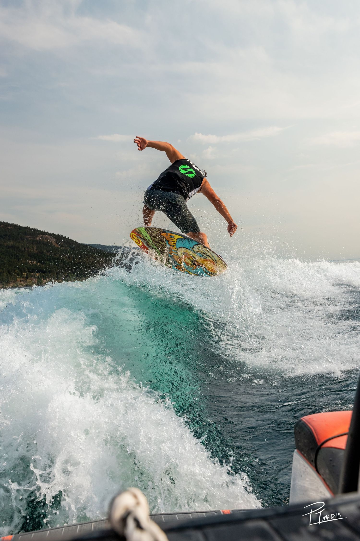 A surfer performs a dynamic aerial maneuver off a wake wave behind a boat on turquoise waters.