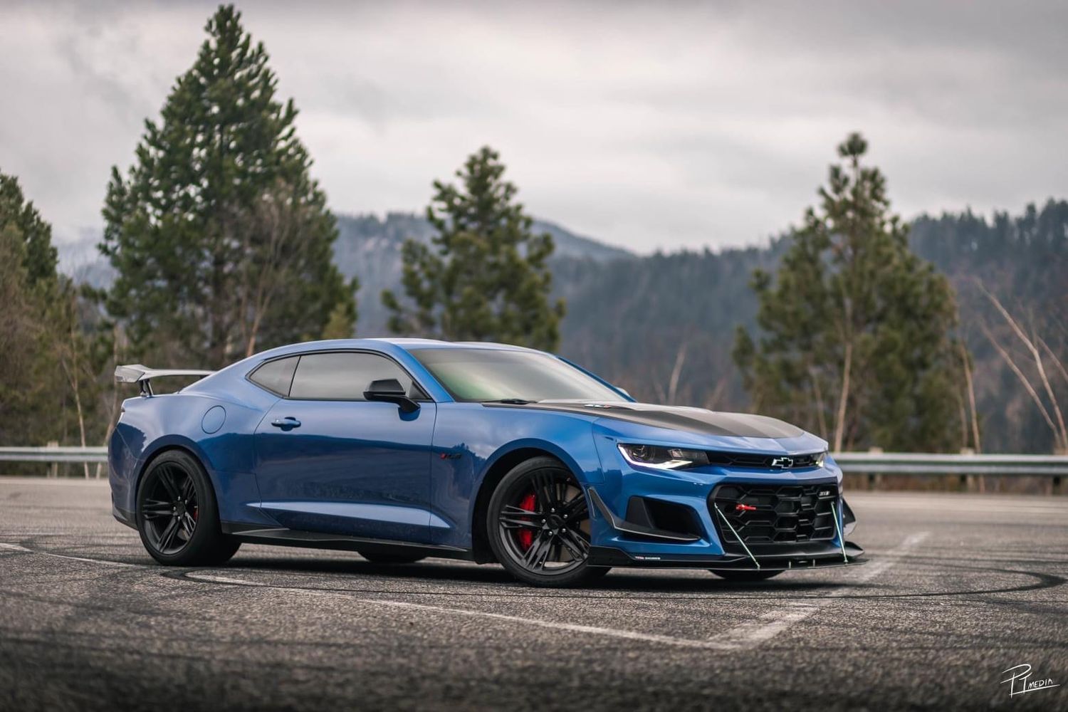 A bright blue Chevrolet Camaro ZL1 sports car parked in front of mountains and evergreen trees on a cloudy day.