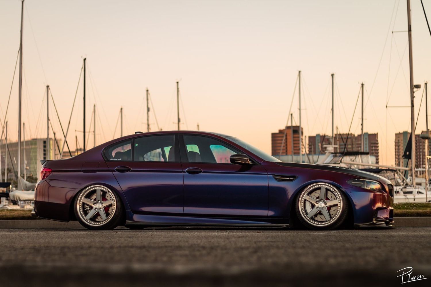 A sleek lowered sports car with chrome wheels parked at a marina during sunset.