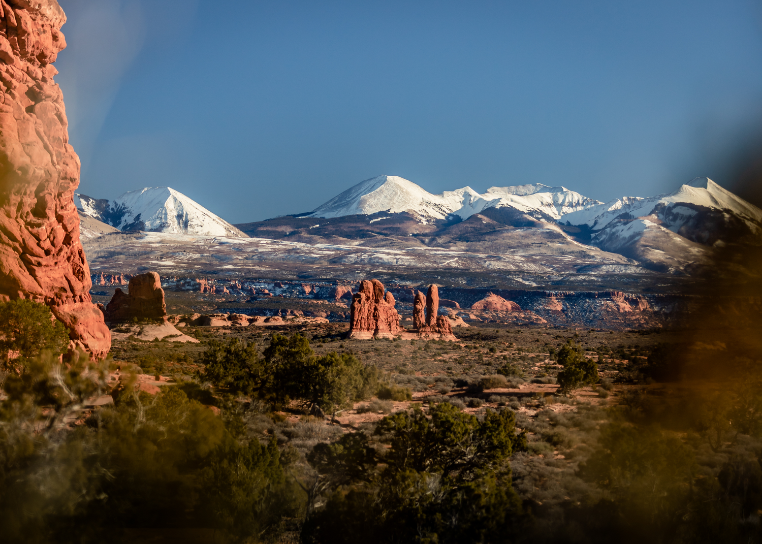 Snow-capped mountains rise behind red rock formations in Arches National Park, Utah.